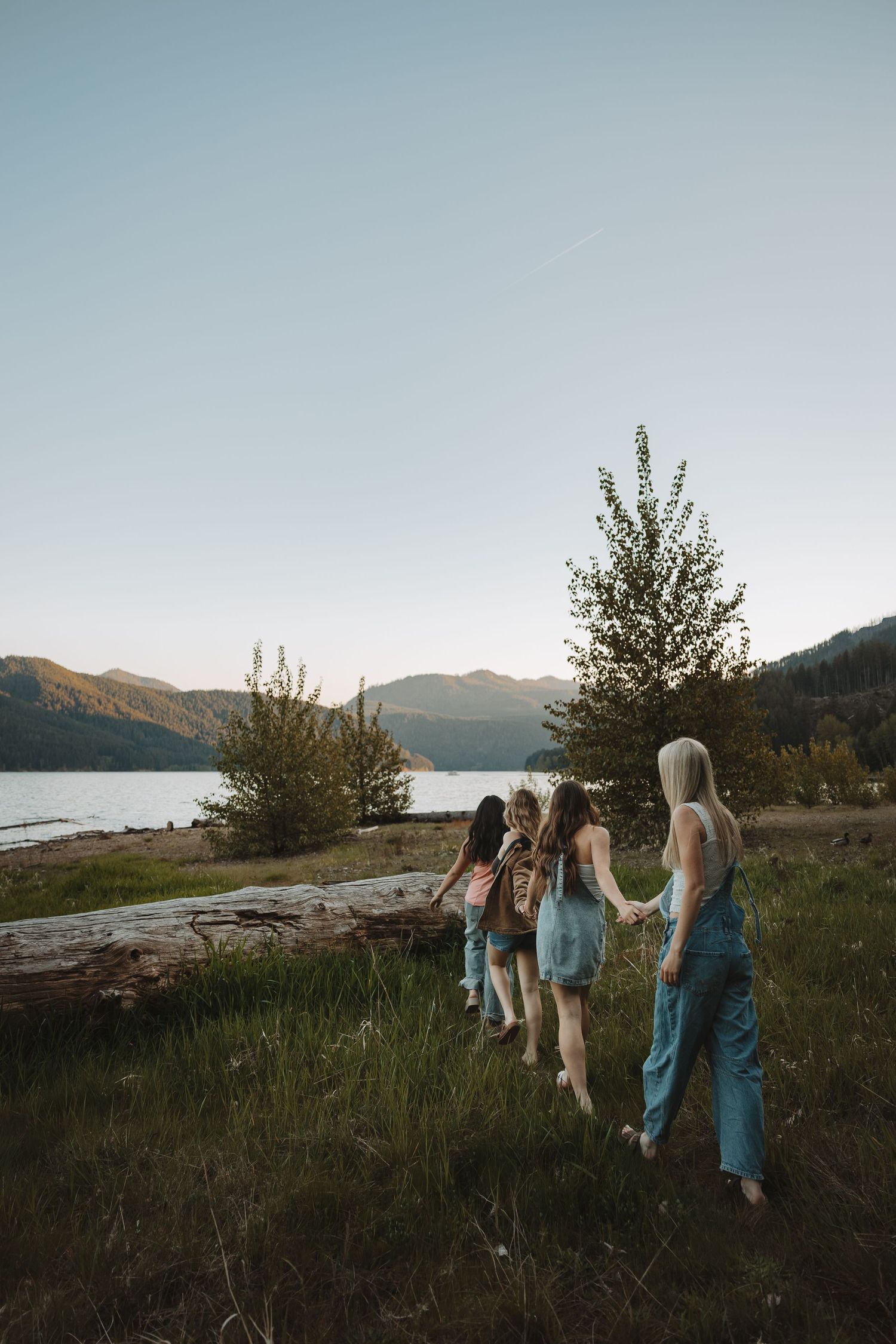 Four young women holding hands walking toward a scenic mountain lake at sunset.