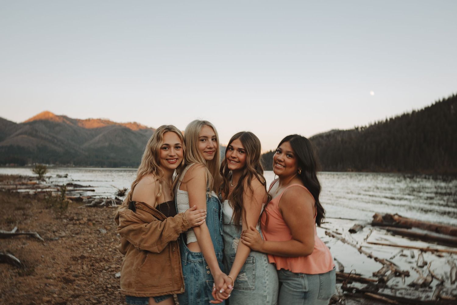 Four young women posing together by a lake with sunlit mountains at dusk.