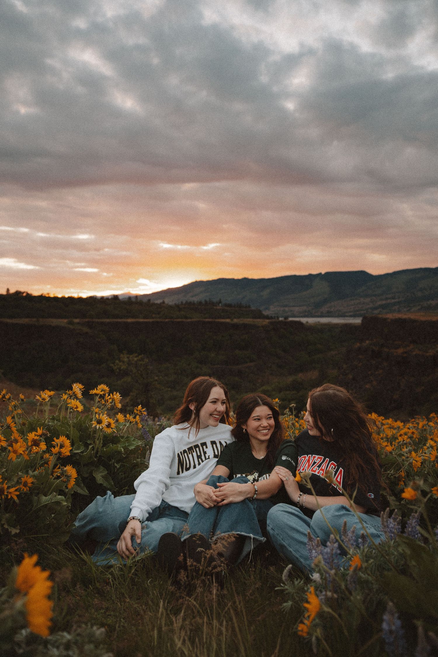 Three young women sitting together in a wildflower field, laughing and enjoying a colorful sunset backdrop.