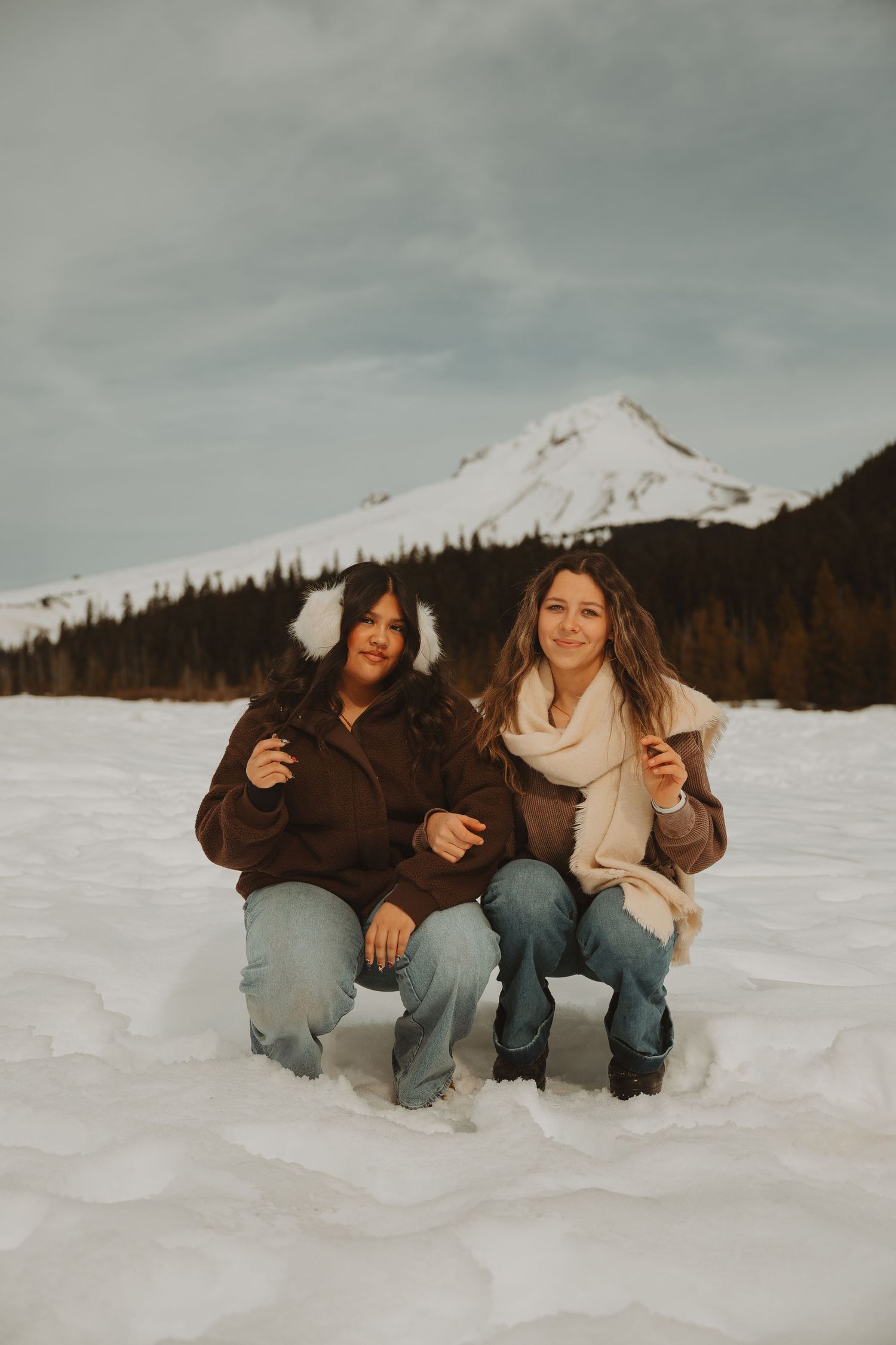 Two women crouch together smiling in snowy alpine setting with mountain and pine trees in background.