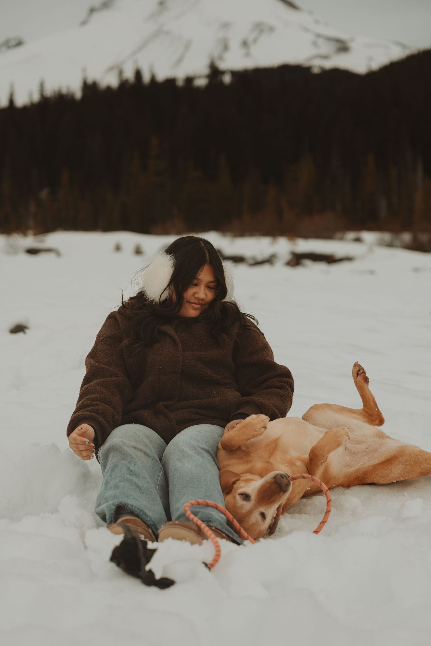 Woman sitting in snow with golden retriever dog in winter mountain landscape.