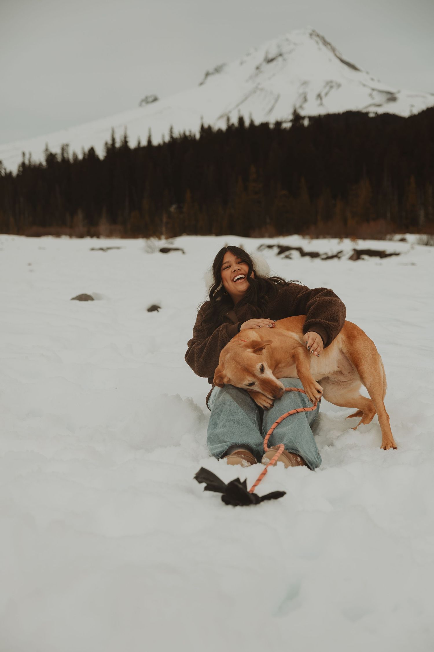 Woman laughing while cuddling golden dog in snowy mountain landscape on cloudy day.