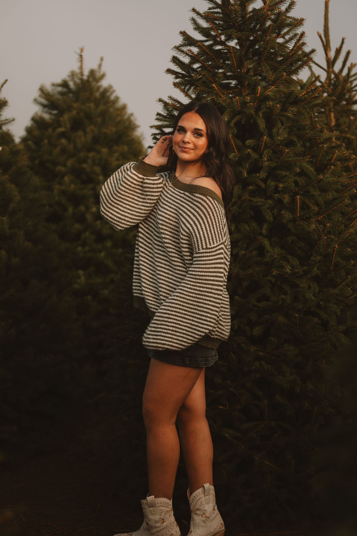 Person in striped sweater poses casually against evergreen trees during golden hour photo shoot.