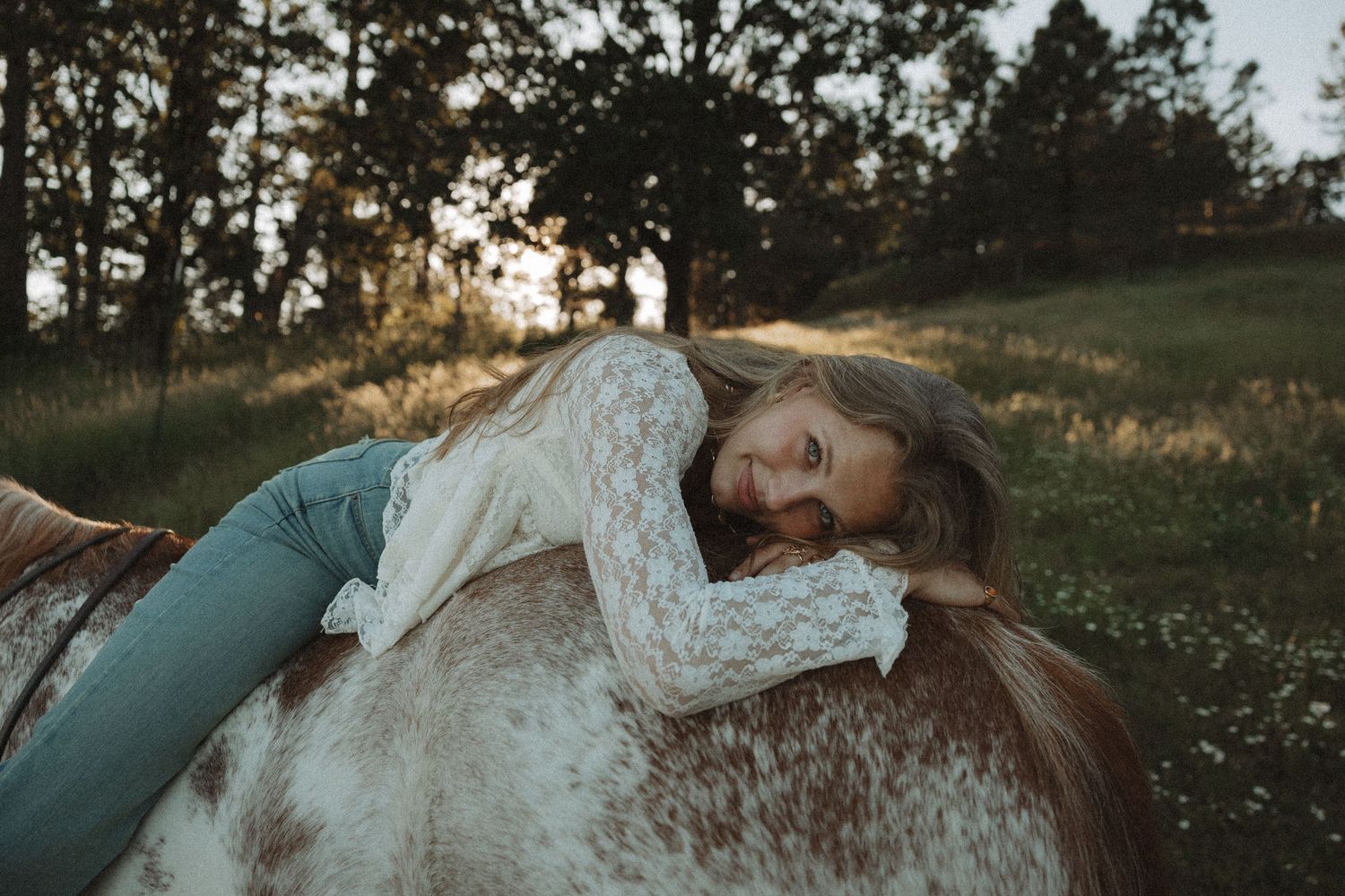Young woman in lace top resting on a paint horse in a sunlit field at golden hour.