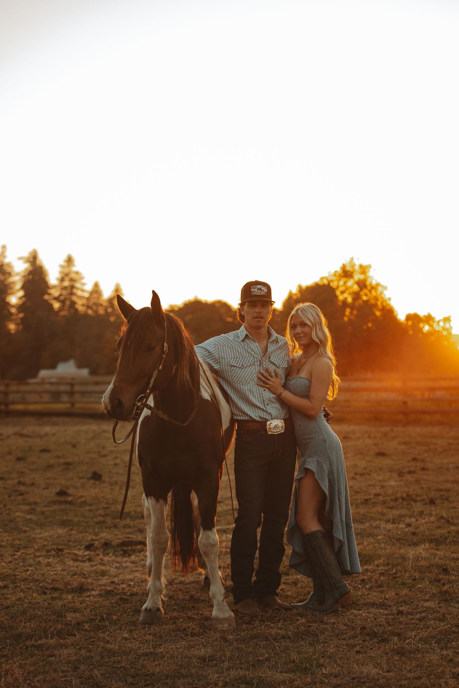 A couple poses with a horse at golden hour sunset on a ranch, woman in blue dress and man in western attire.