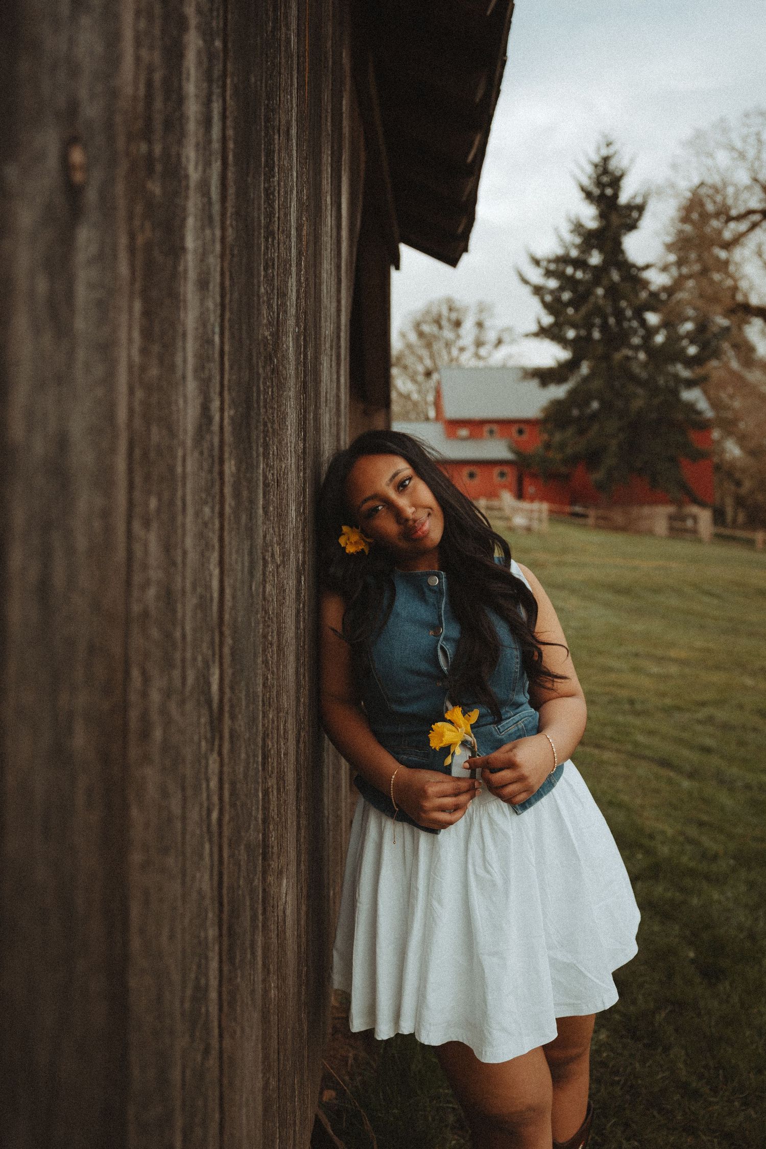 A person in a denim top and white skirt leans against a rustic wooden post in a rural setting.