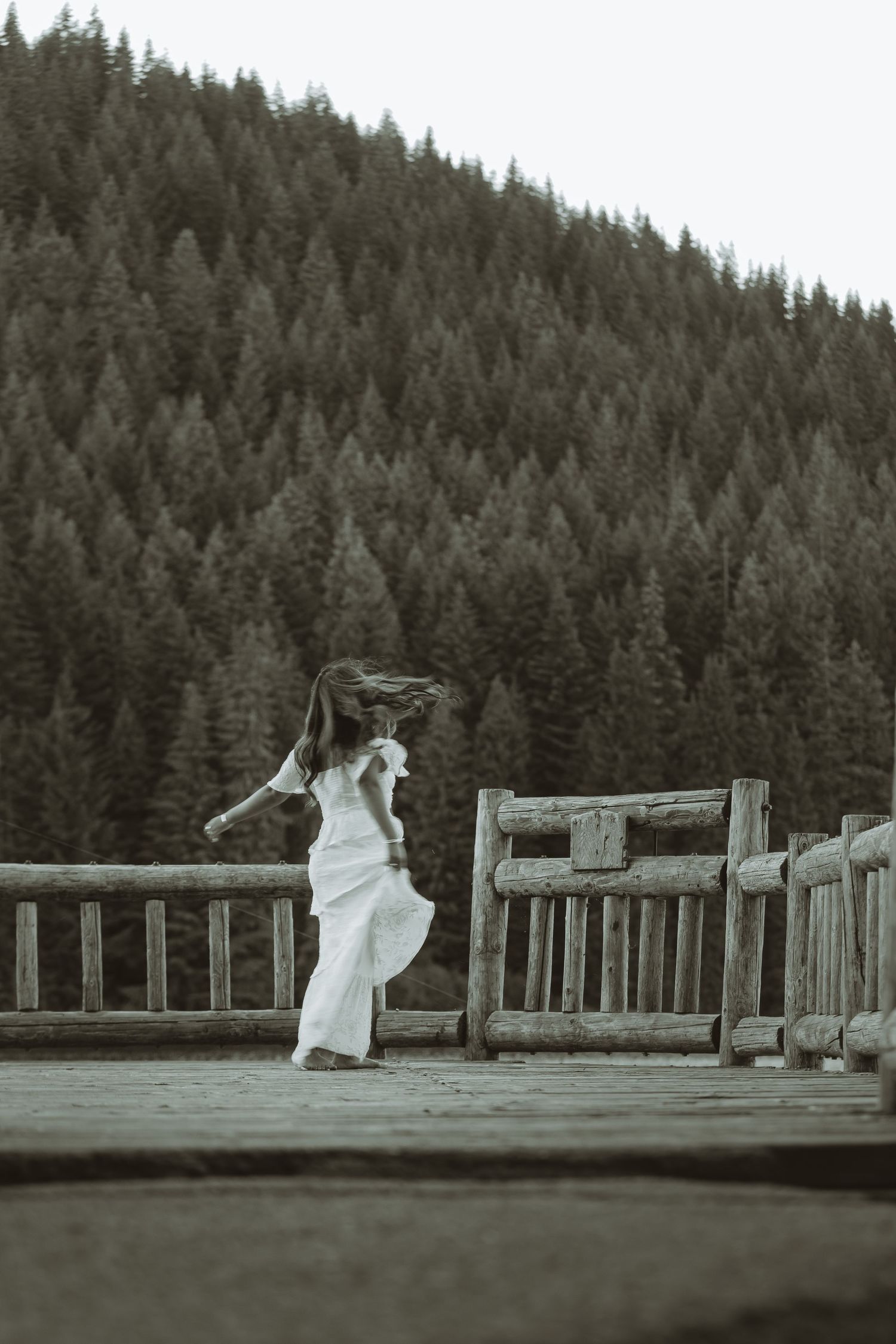 A person in a flowing white dress dances on a wooden bridge with evergreen forest mountains in the background.