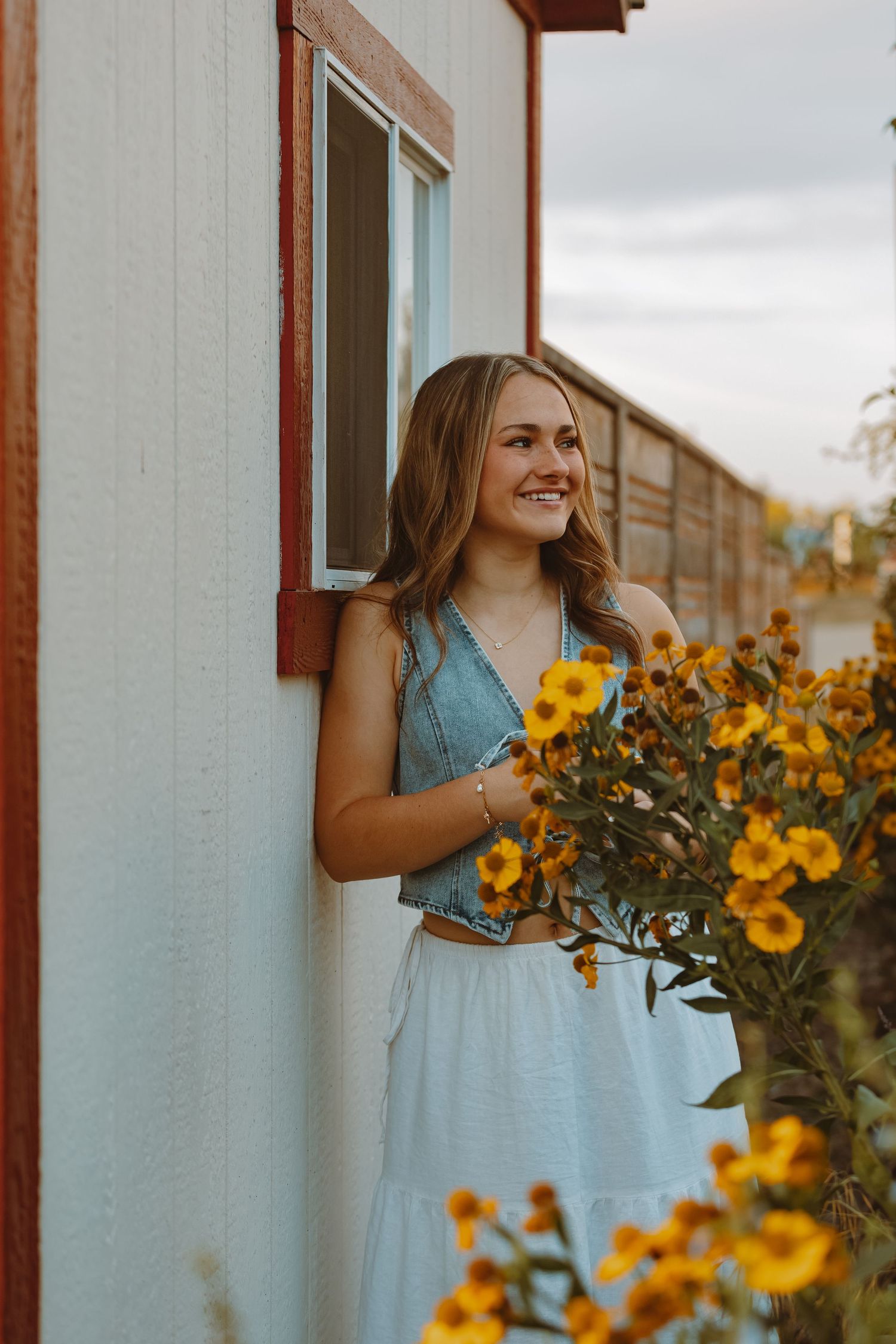 A person in a blue dress stands by a white building with blooming yellow sunflowers in the garden.