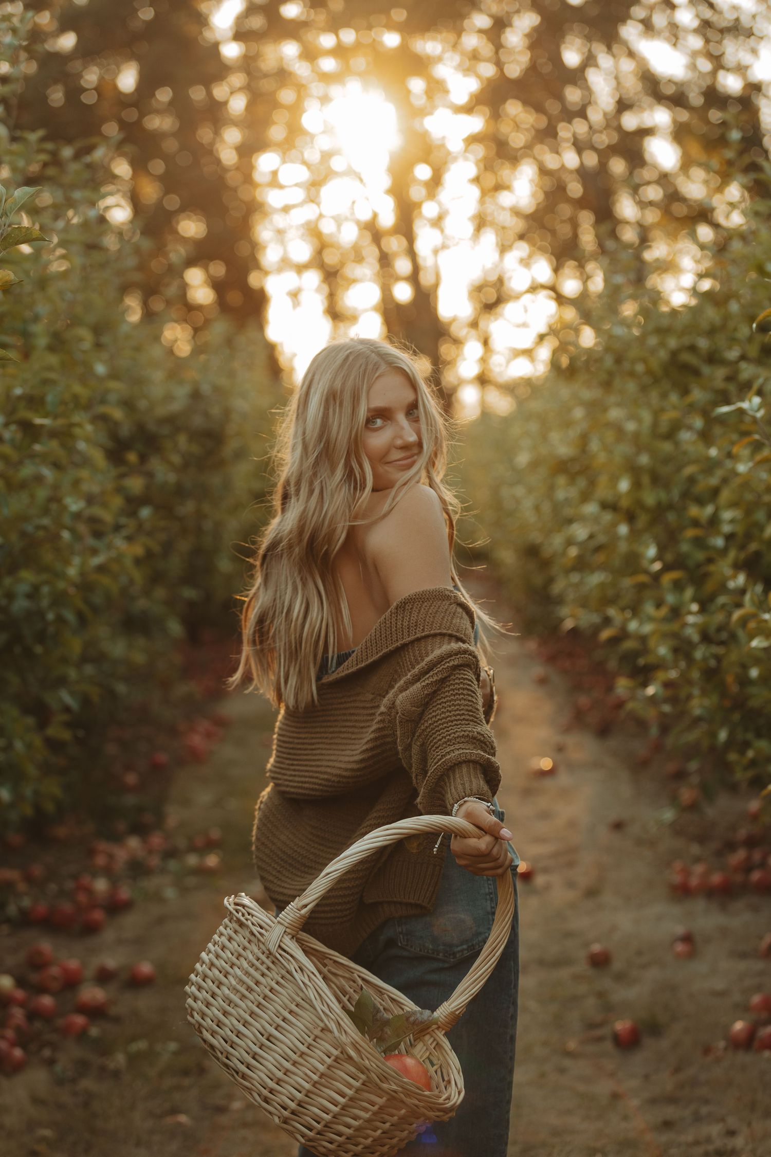 A figure in a brown sweater holds a woven basket while walking through an apple orchard at golden sunset.