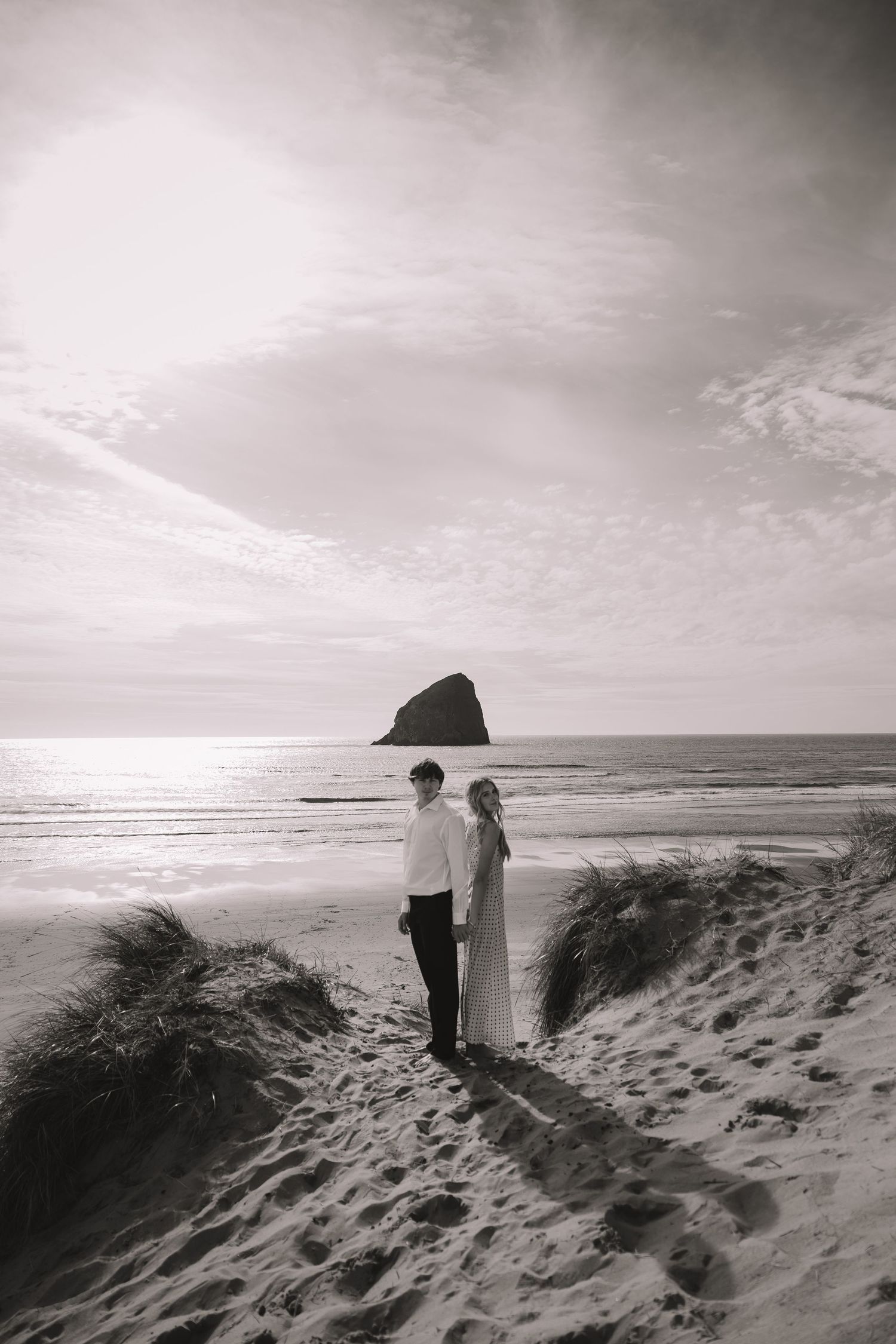 Black and white photo of a couple standing on sandy beach dunes with a sea stack rock formation in the background.