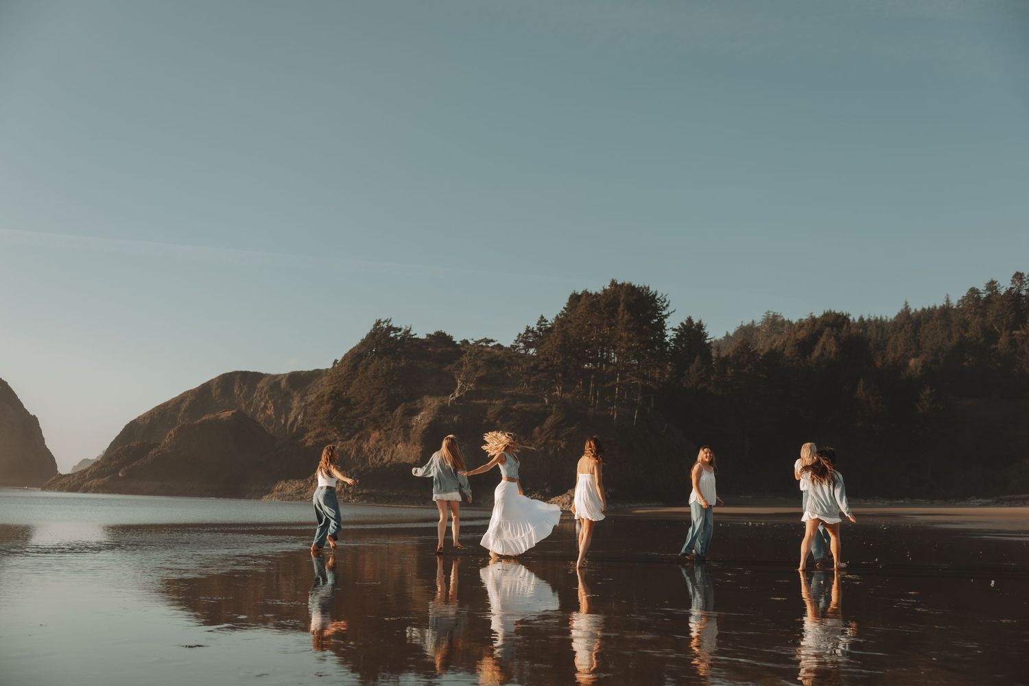 Group of women in white dresses dancing on a reflective beach with rocky cliffs at sunset.