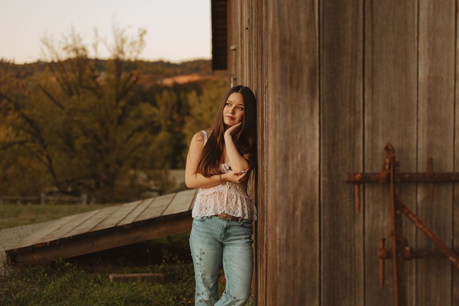 Young woman in lace top and jeans leaning against rustic barn wall during golden hour sunset.