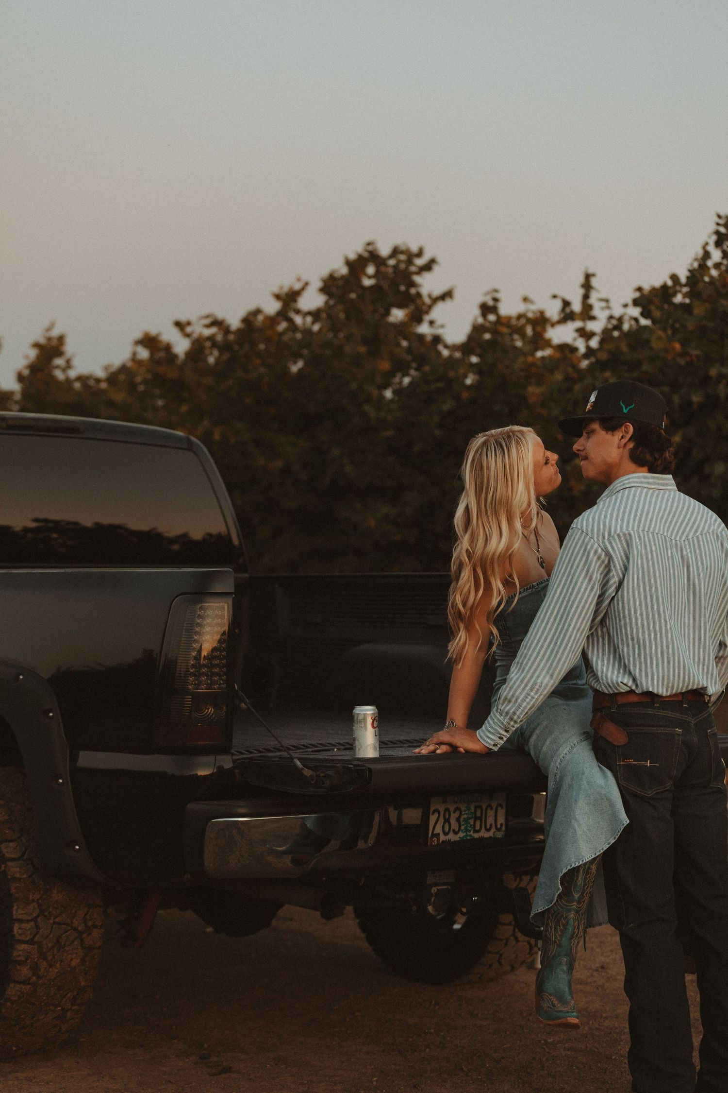 Couple sharing a kiss while sitting on a truck tailgate at dusk near an orchard.