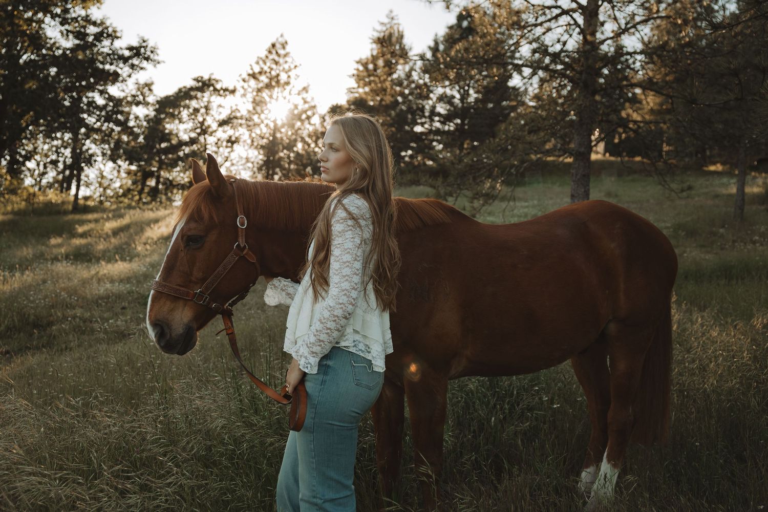 A young blonde woman in a white blouse and jeans stands beside a chestnut horse in a sunlit meadow.