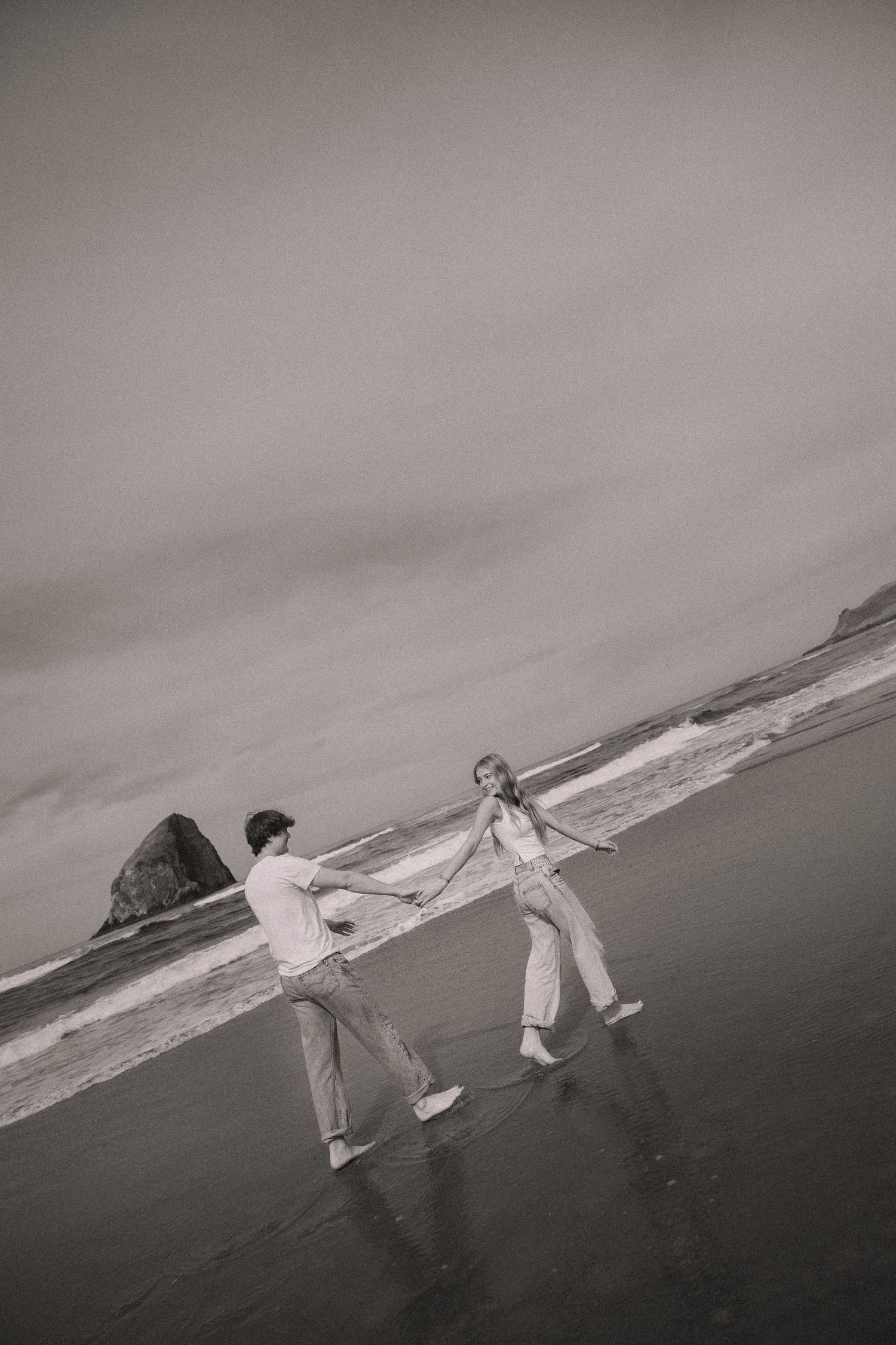 Black and white photo of a playful couple holding hands and dancing on a wet beach with a sea stack in the background.