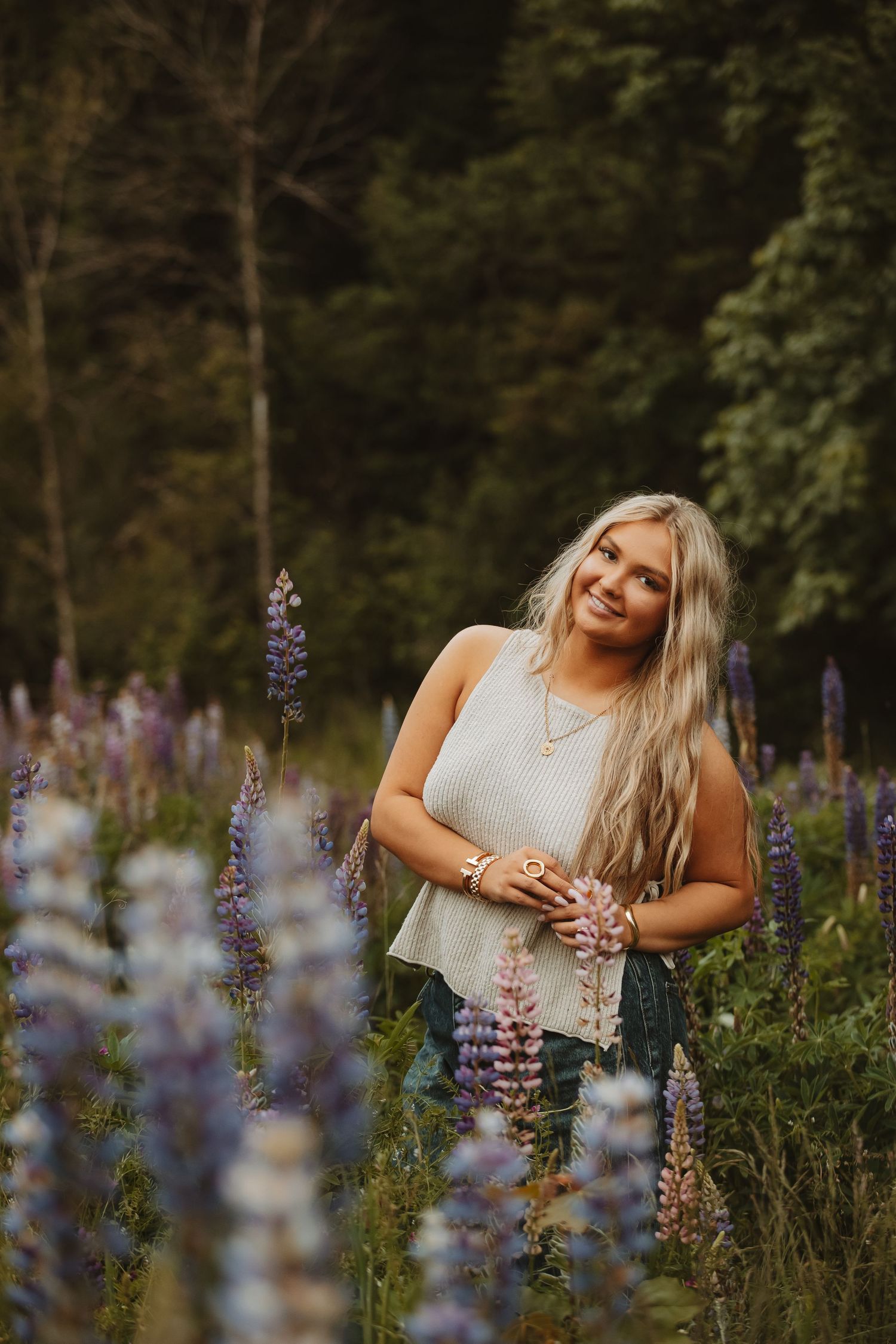 Woman in casual attire poses among purple lupine flowers at sunset.