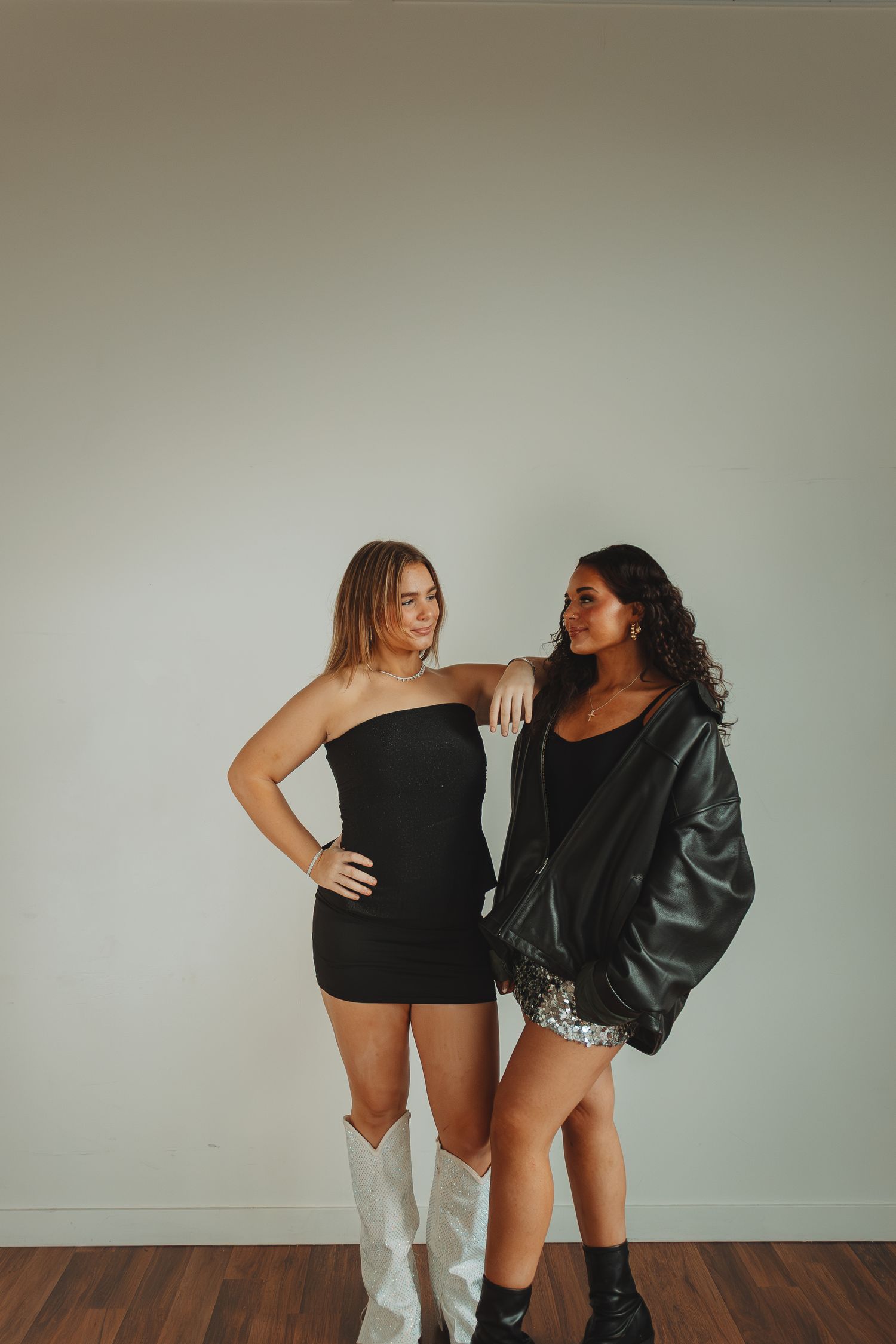 Stylish duo posing in black outfits against minimal white studio backdrop.