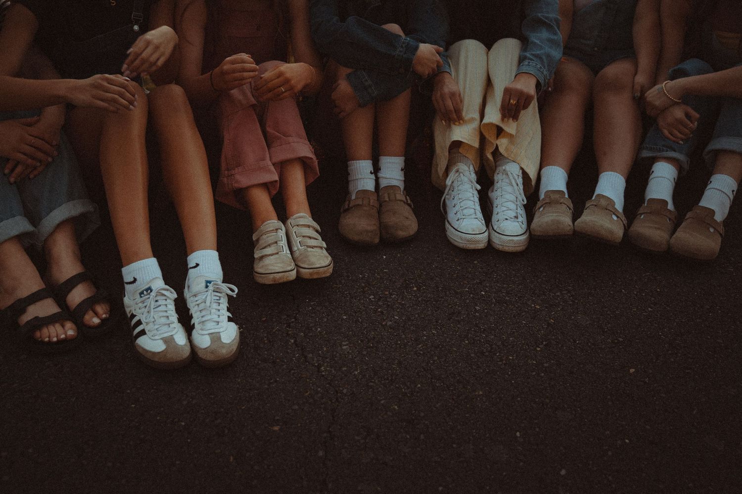 Group of friends sitting together showing off their stylish sneakers and casual footwear on pavement.