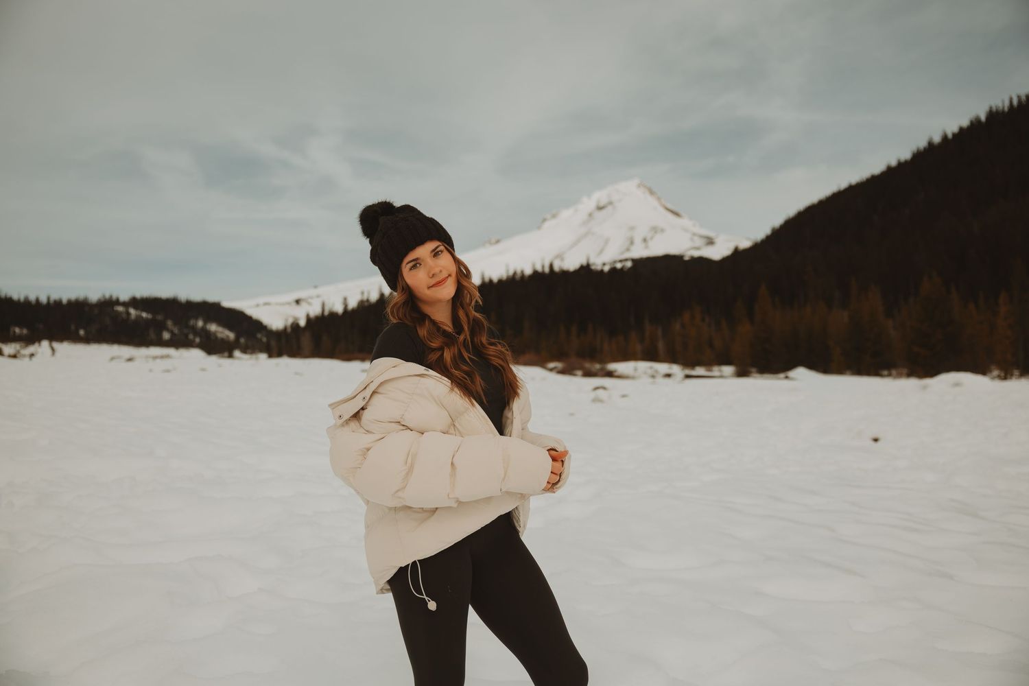 Young woman in white puffer jacket and black beanie smiling in snowy mountain landscape.