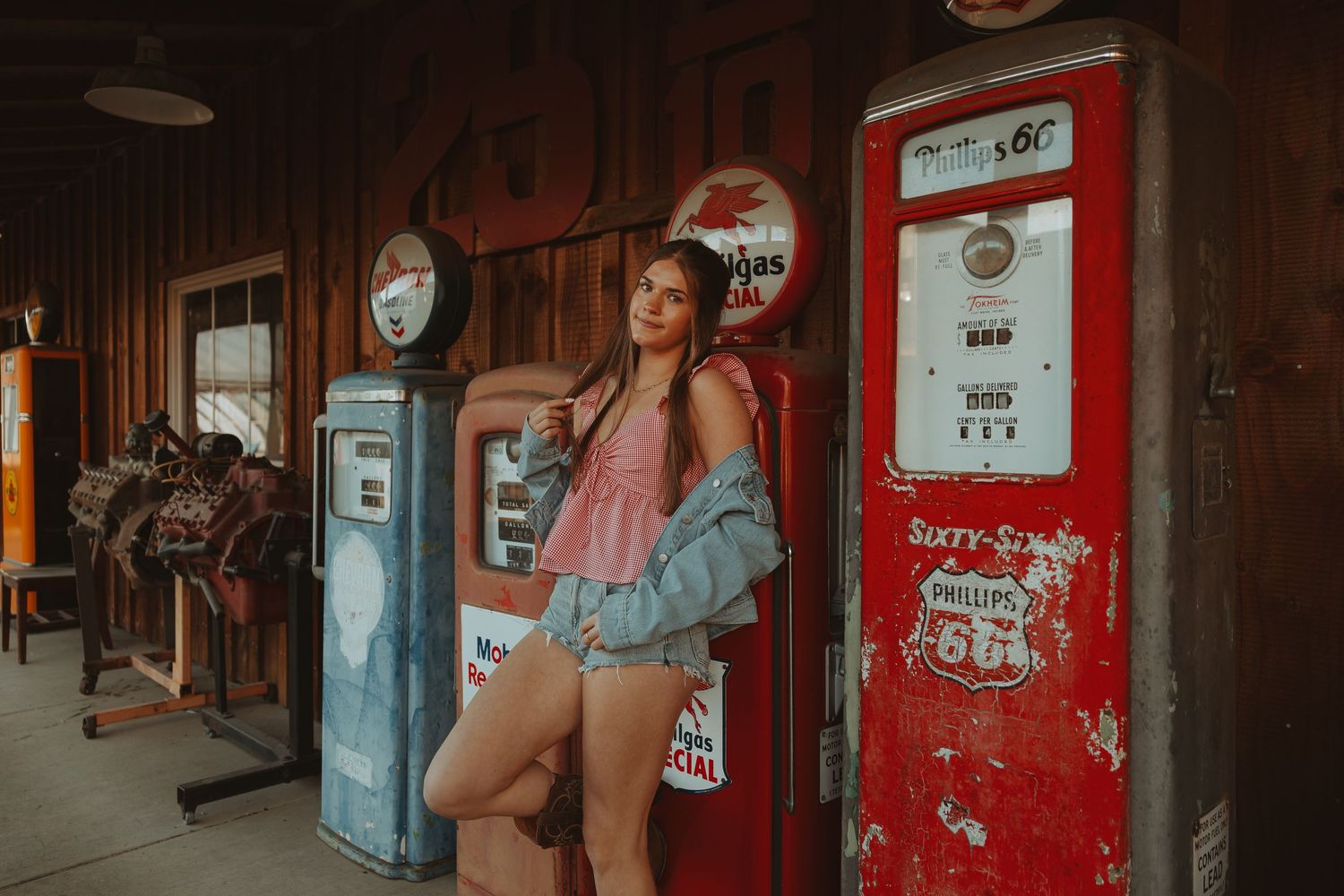 Young woman in denim jacket poses beside vintage red gas pumps at a rustic retro Route 66 station.