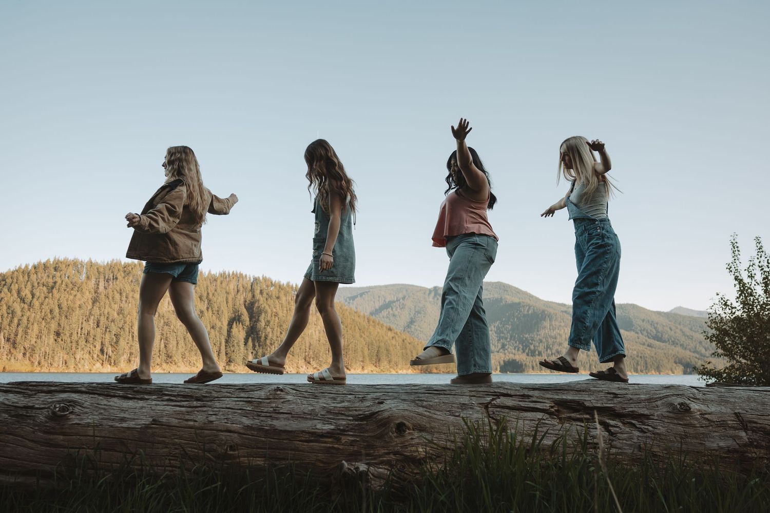Four girls balance and walk along a large fallen log near a scenic mountain lake surrounded by evergreen trees.