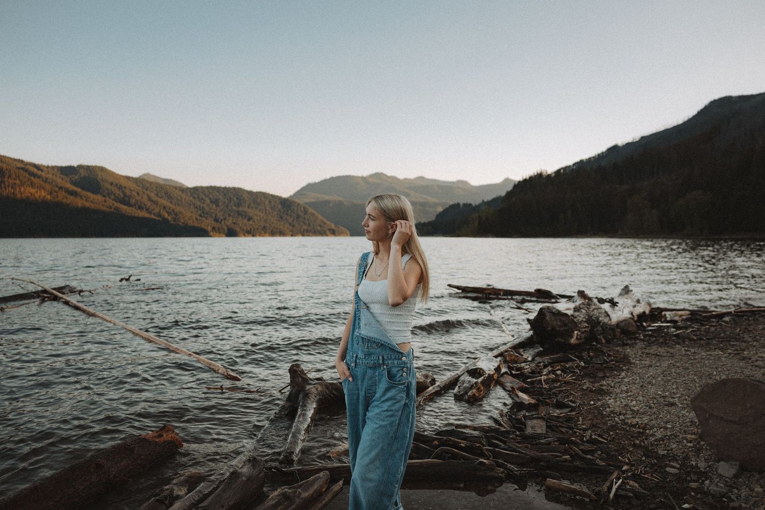 Blonde woman in blue overalls standing by a serene mountain lake with driftwood on the shore.