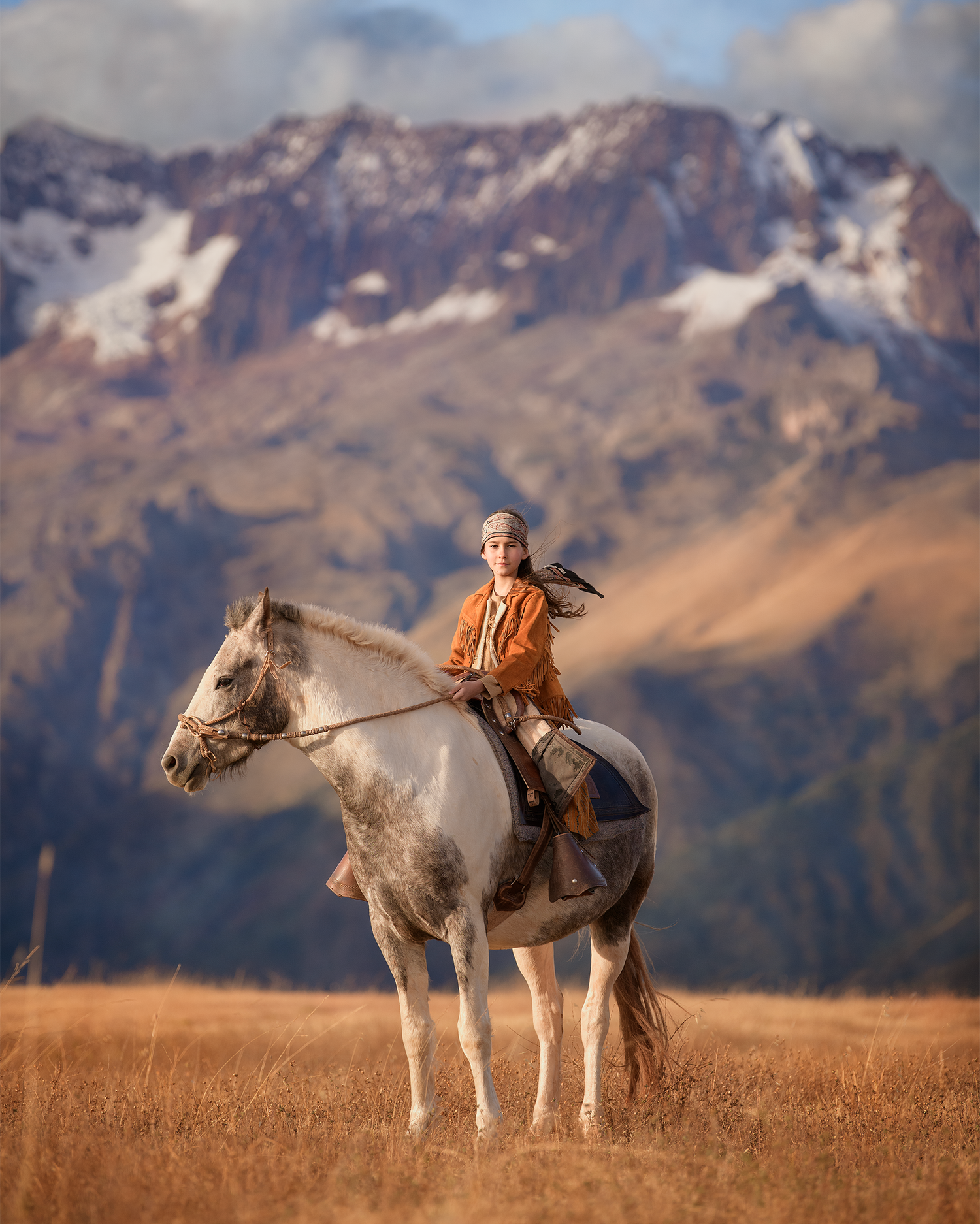 A rider on a white horse stands against a backdrop of majestic mountain peaks in golden afternoon light.