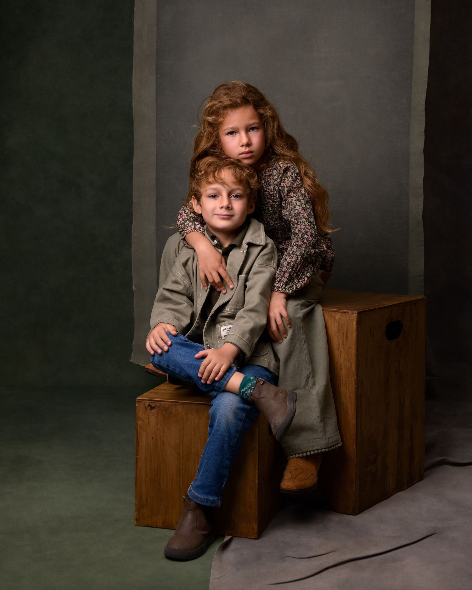 Two red-haired children pose together on a wooden box in a studio portrait, wearing stylish autumn outfits and boots.