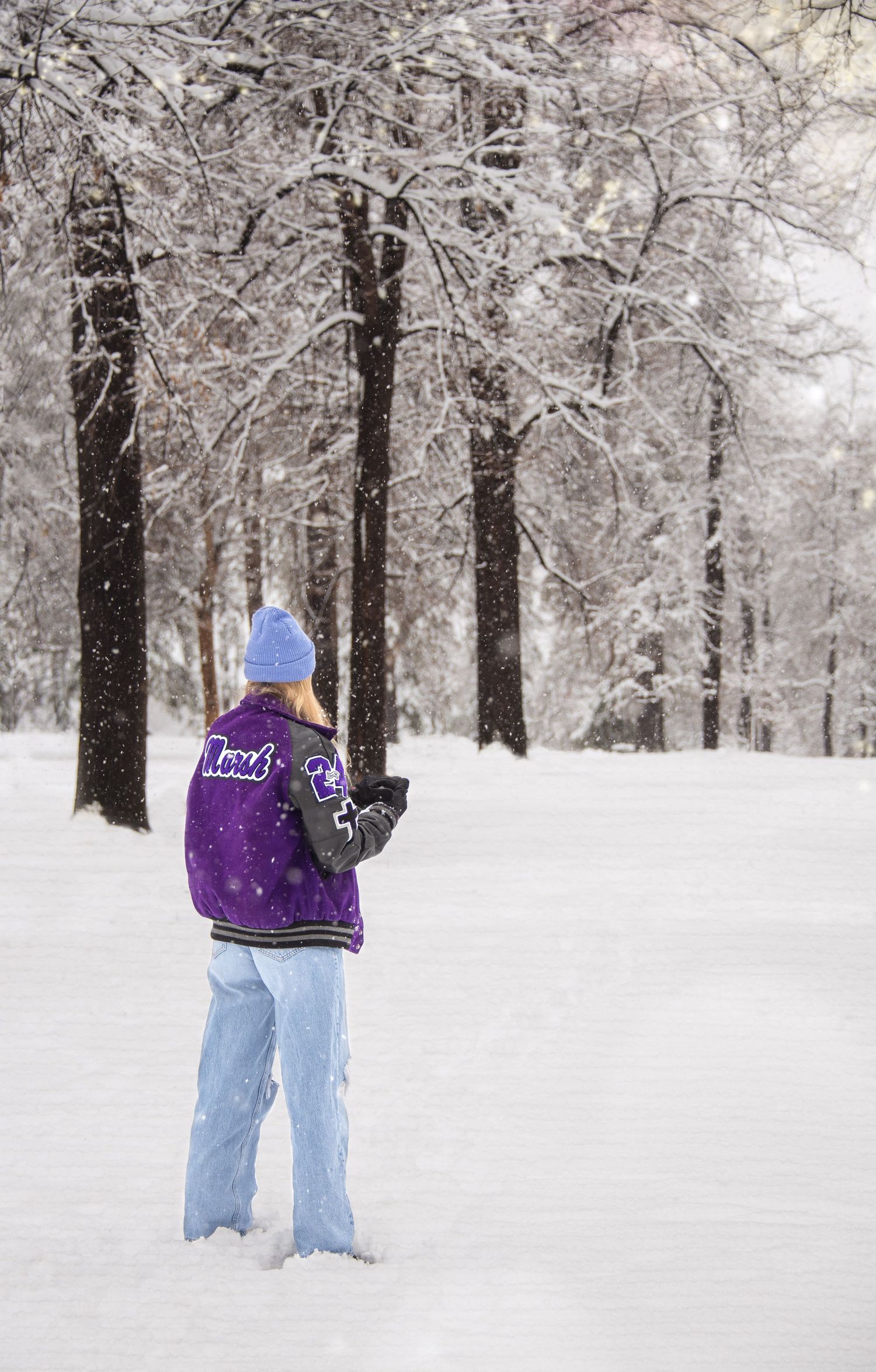 Teenager in purple jacket and blue hat standing in snowy winter forest.