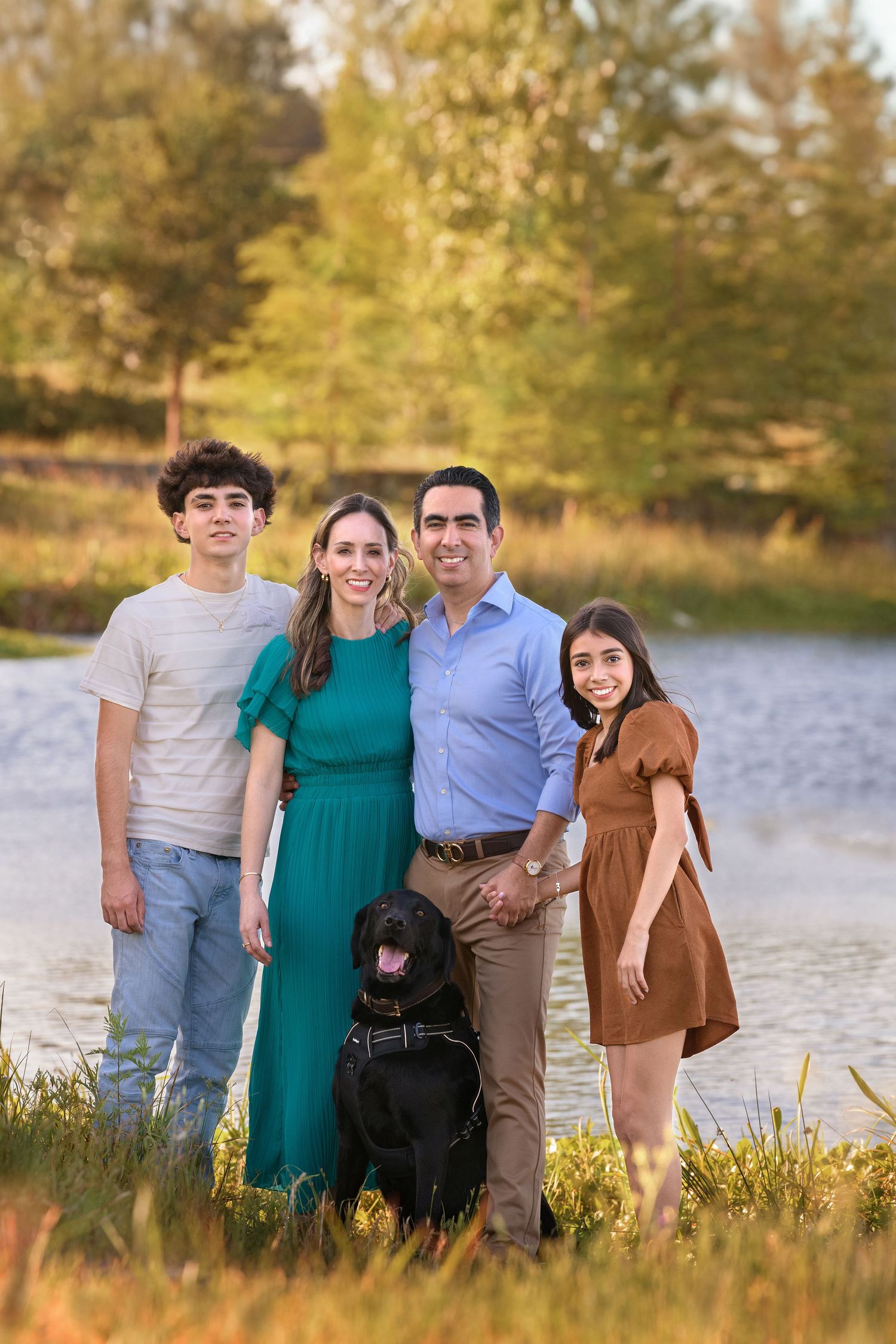 A family of four and their black dog pose beside a lake during golden hour in an autumn setting.