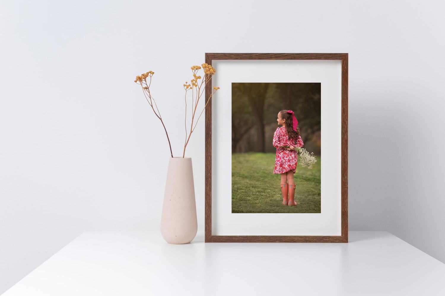 Framed portrait photograph of little girl displayed on white surface with minimal vase and dried flowers.