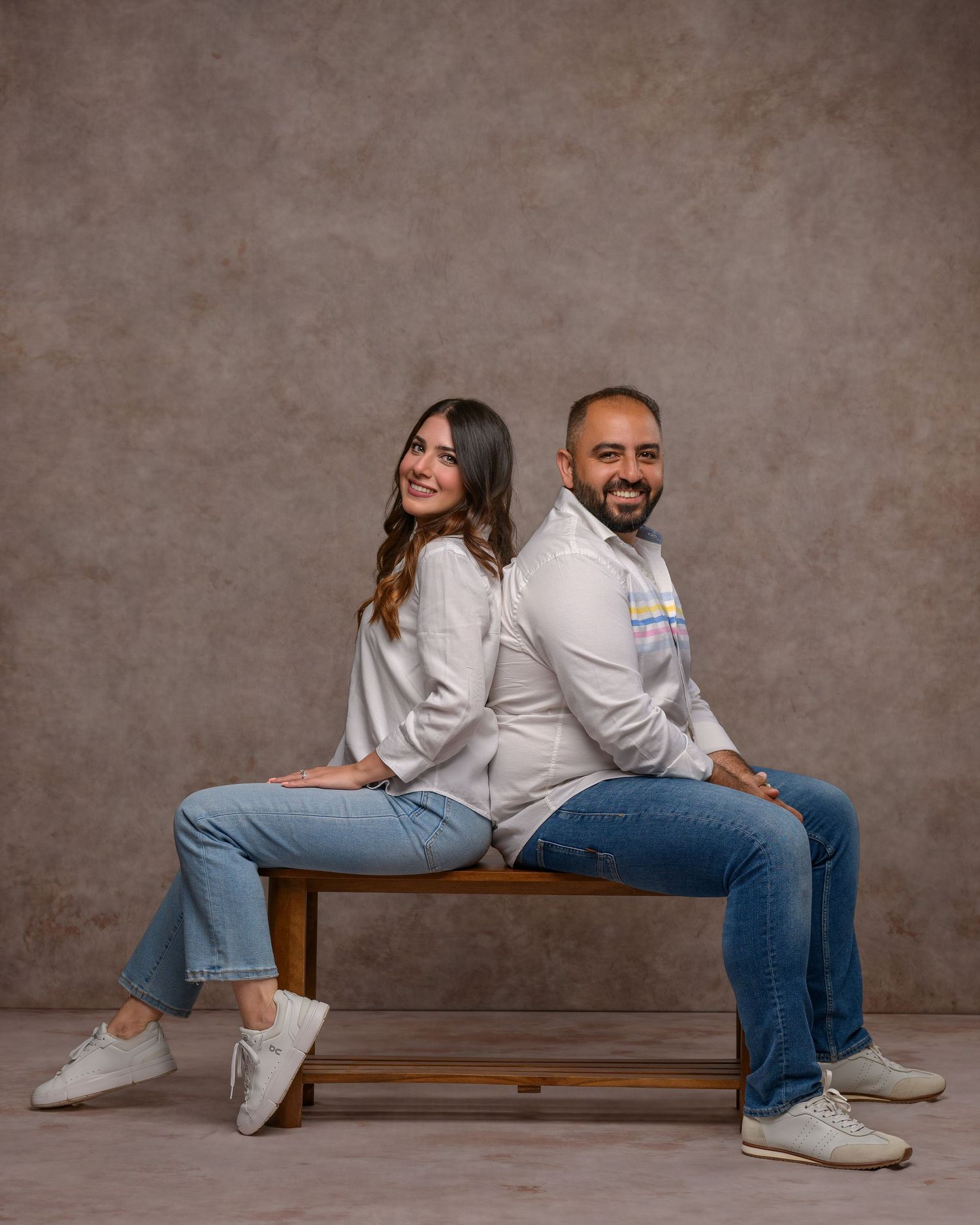 A smiling man and woman sit back-to-back on a wooden bench, wearing white shirts and jeans against a gray backdrop.