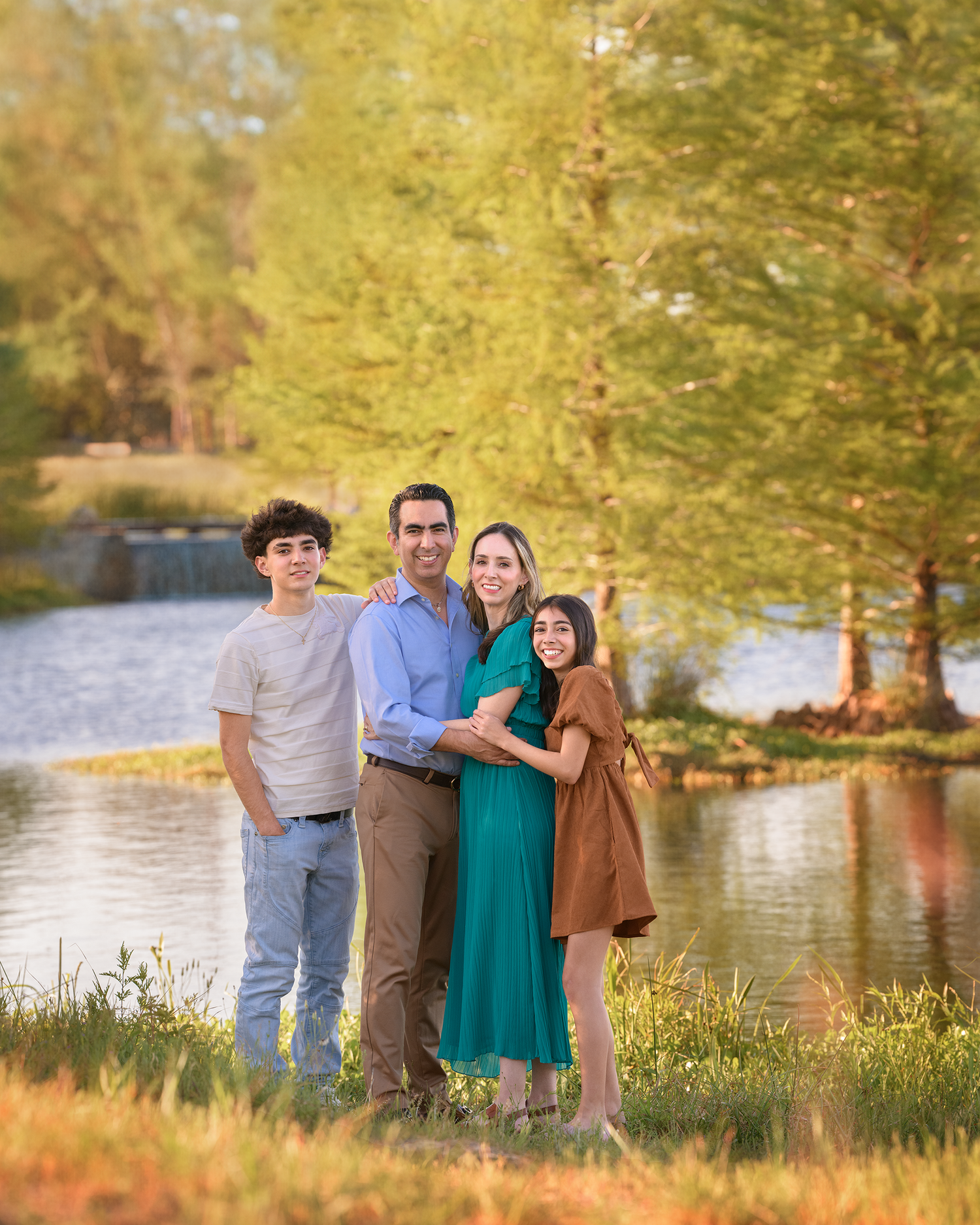 A family stands together on a grassy bank beside a lake during golden hour with autumn trees in the background.