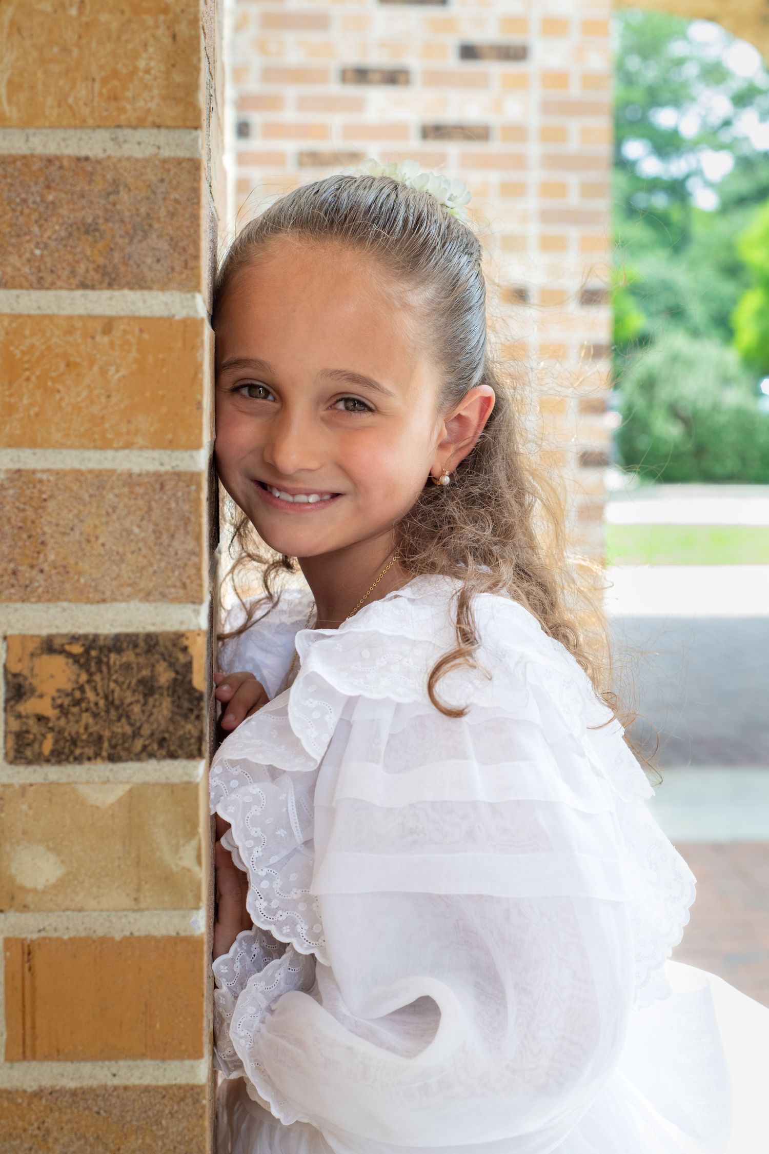 Young girl in white ruffled dress smiling beside brick column.