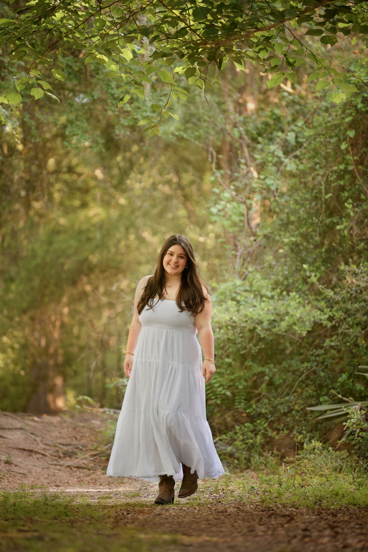 A person in a flowing white dress walks along a peaceful forest path surrounded by lush green foliage.