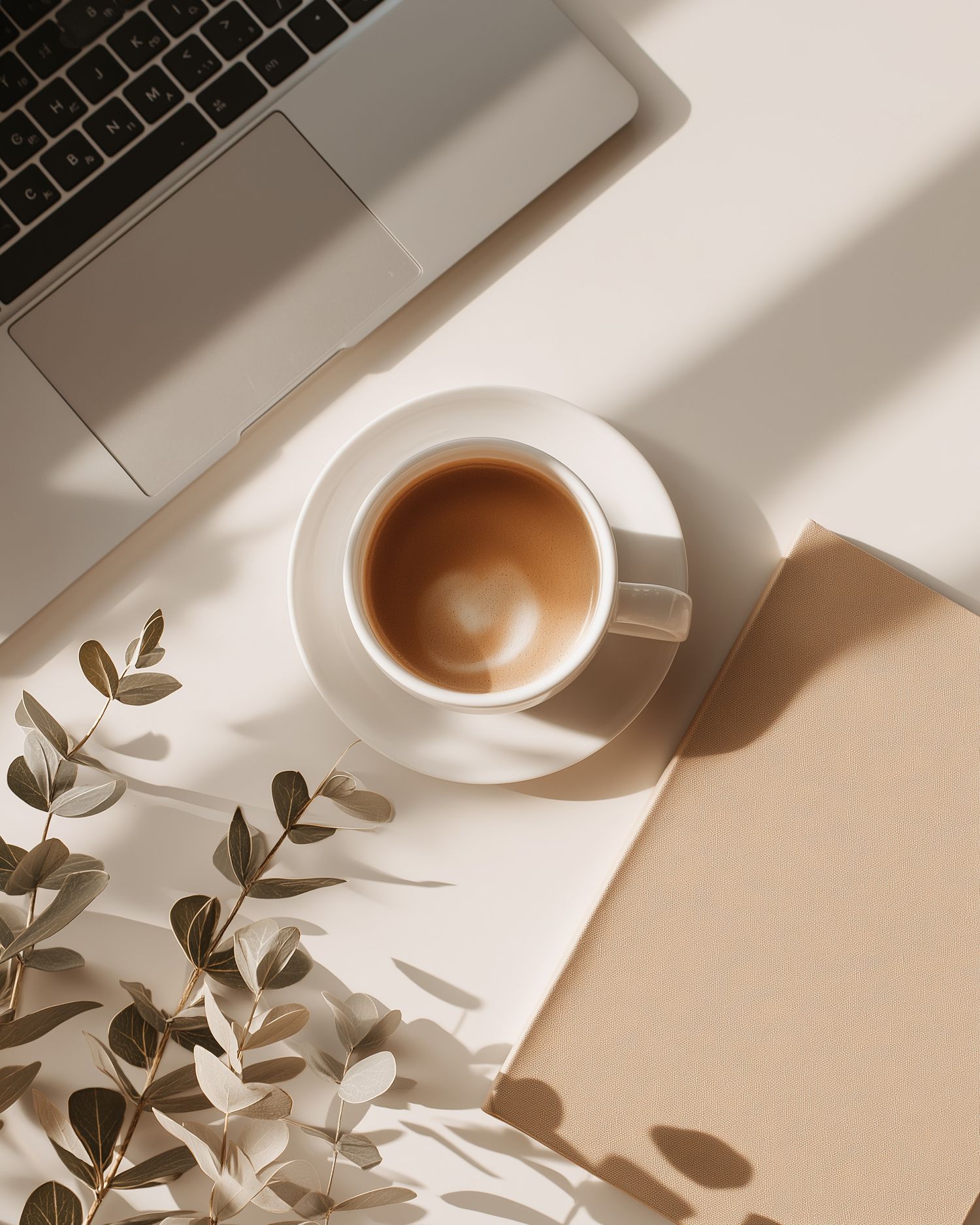 Minimalist flatlay composition of coffee cup and plant leaves casting shadows on white desk with laptop keyboard.