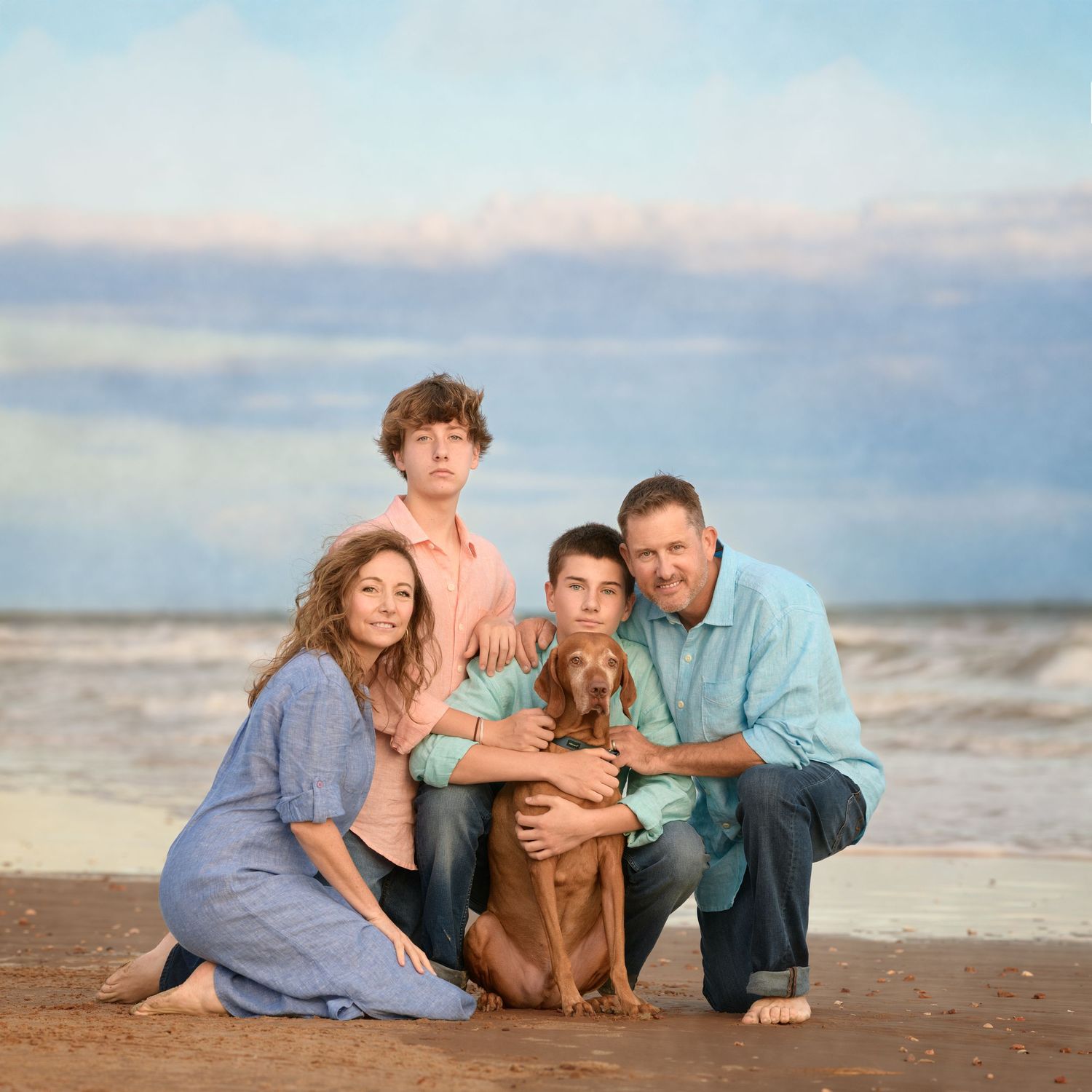 A family poses together with their brown dog on a beach at sunset with cloudy skies in the background.