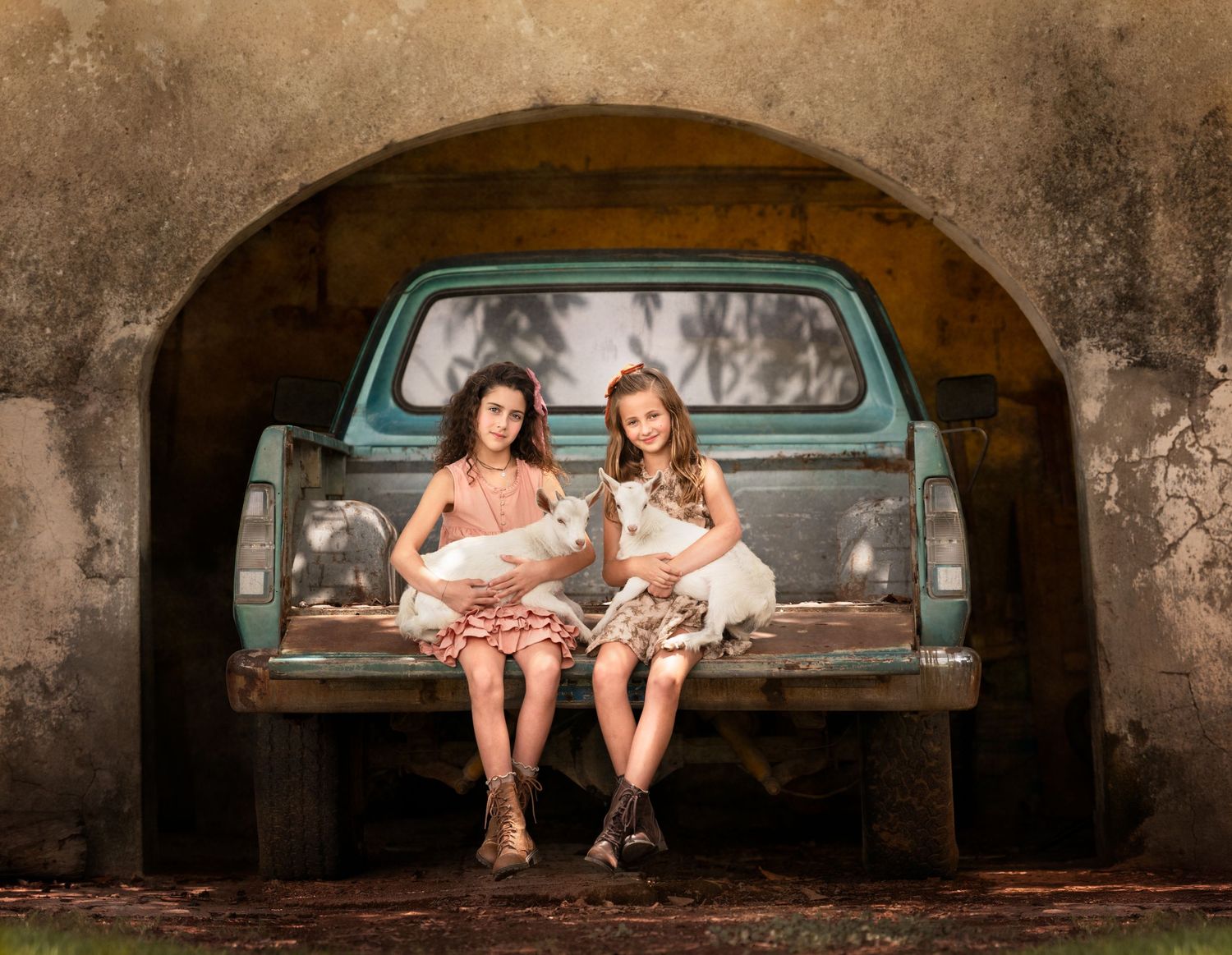 Two girls sitting in vintage turquoise pickup truck bed under rustic stone archway.