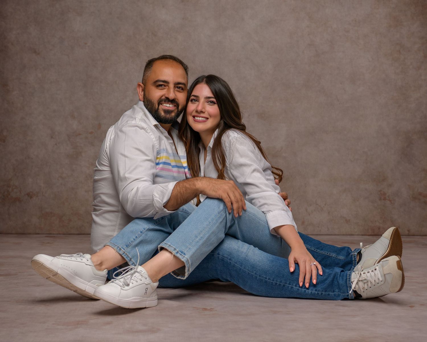 A smiling couple in white shirts and jeans sitting together on the floor in a studio portrait setting.