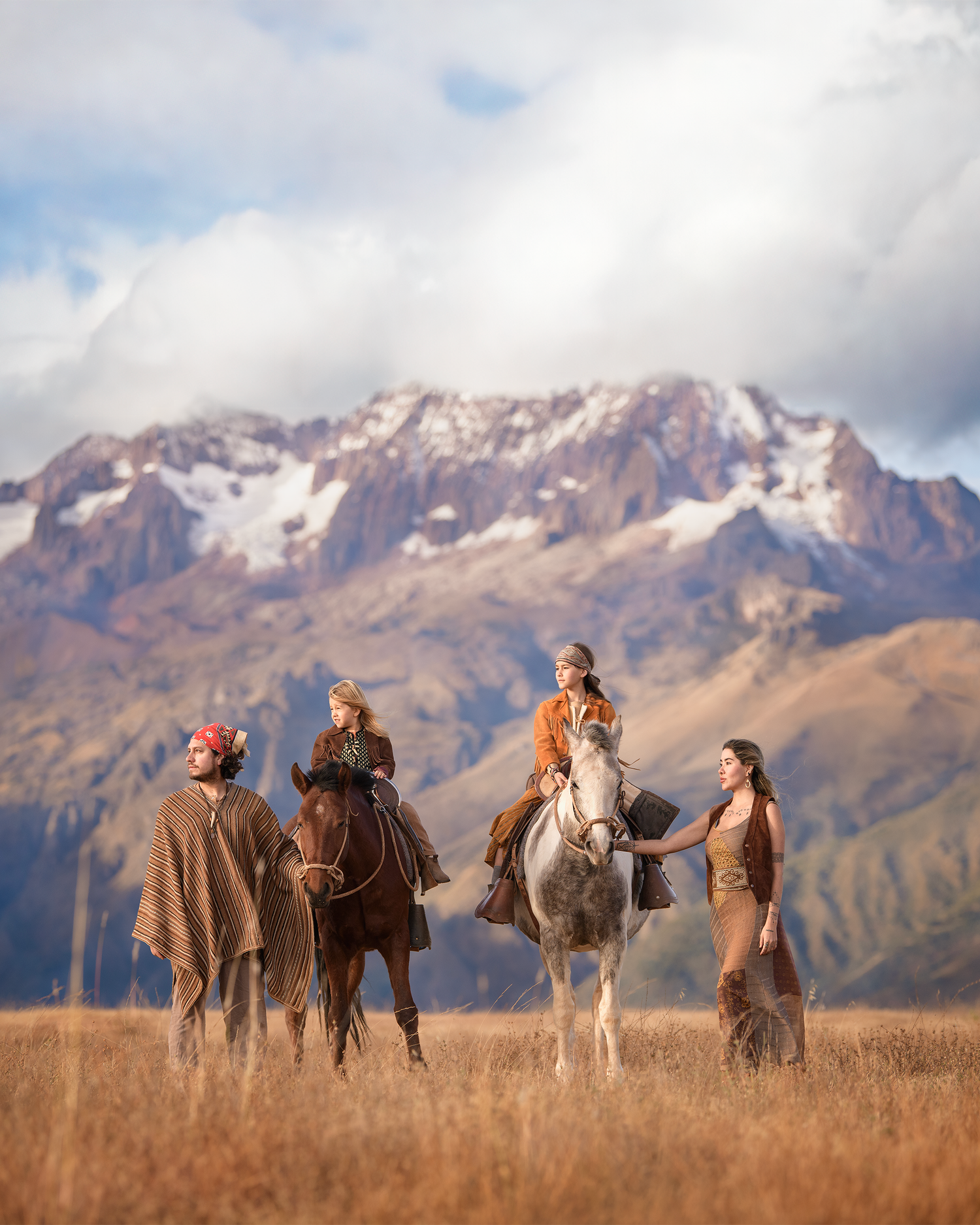Horseback riders gather against a dramatic mountain backdrop with snow-capped peaks and golden grasslands in the foreground.