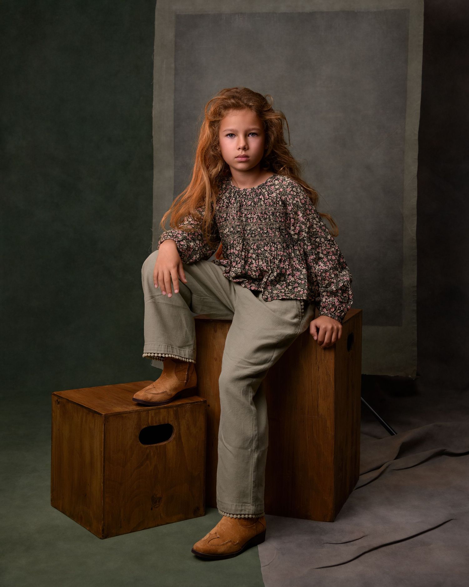 A young girl with long red hair sits on a wooden box, wearing a floral top, khaki pants, and tan boots.