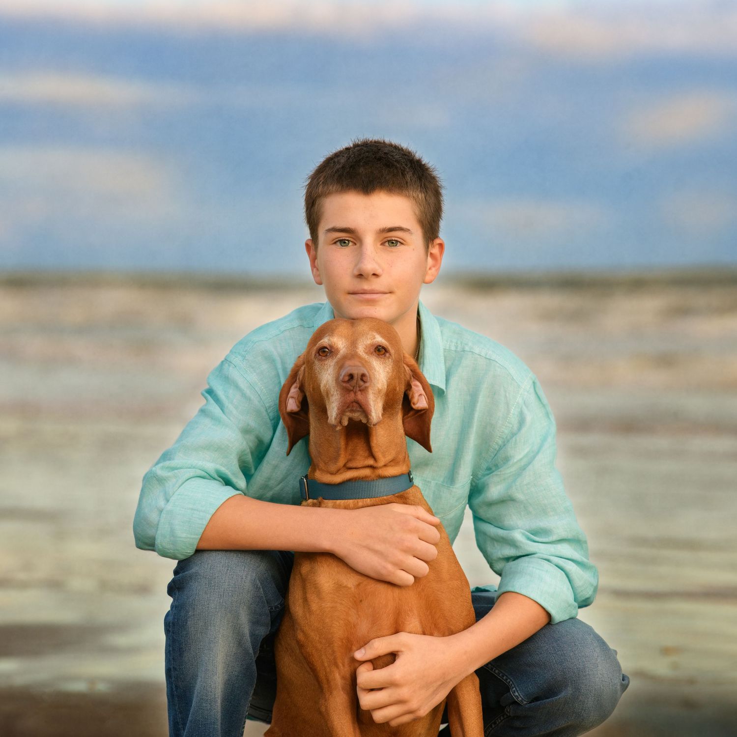 Teenager in turquoise shirt hugging brown Vizsla dog on beach at sunset.