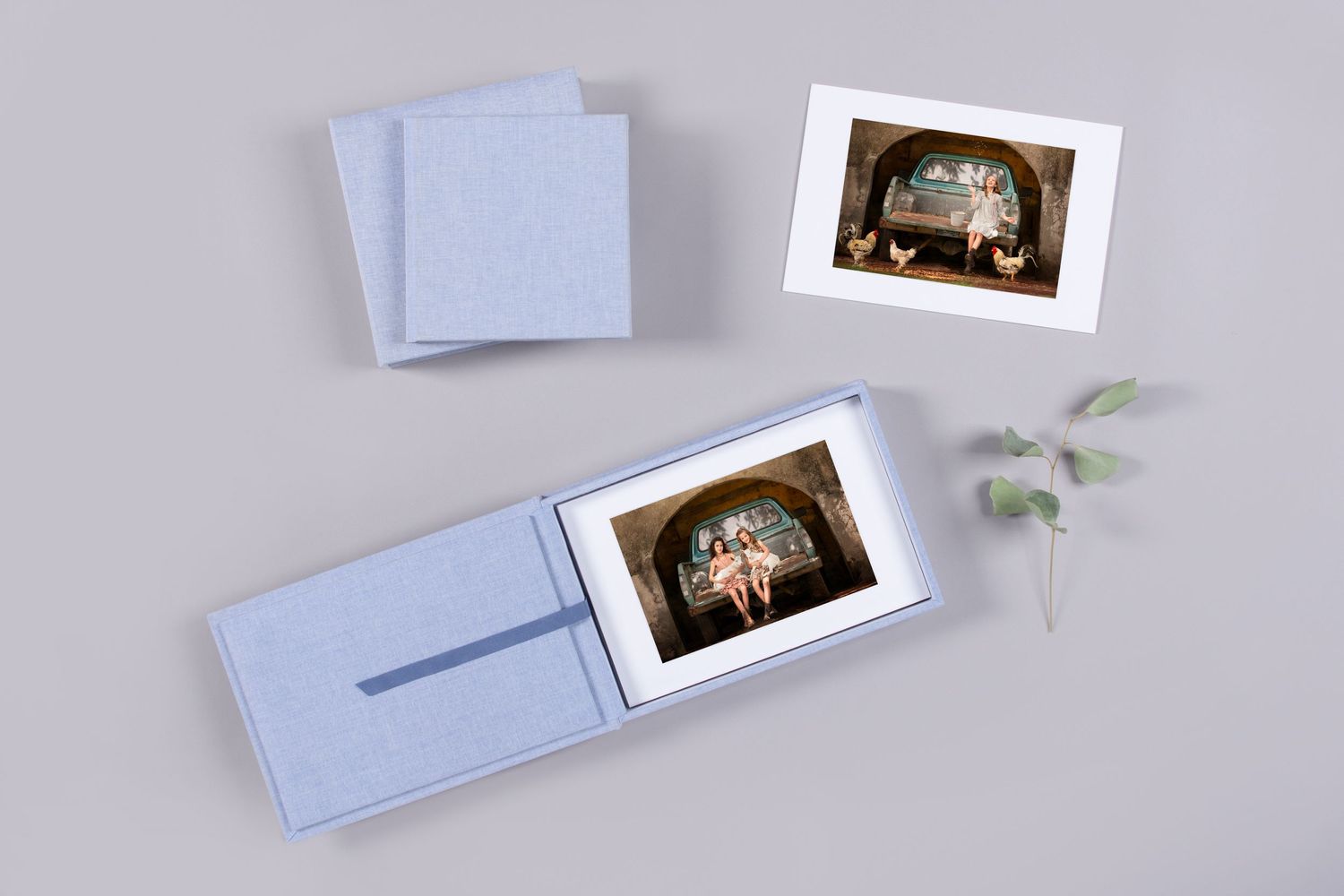 White photo folders and prints displayed with a delicate sprig of greenery on a light background.