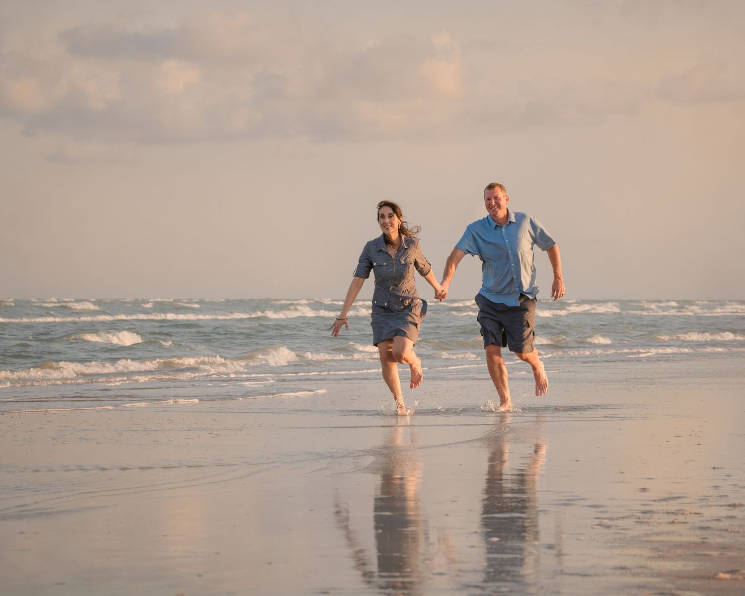 Couple holding hands running barefoot on a sandy beach with ocean waves in the background at sunset.