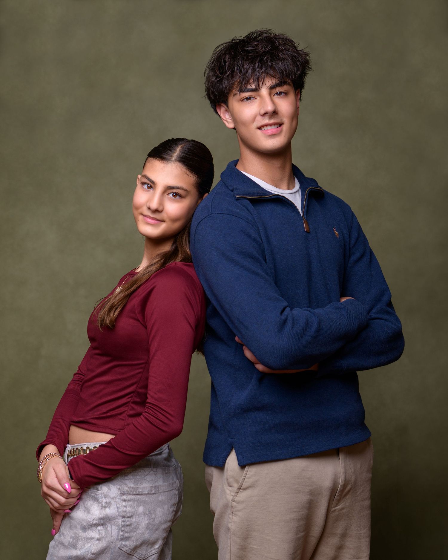 Studio portrait of two young people in casual attire standing back to back against an olive green background.