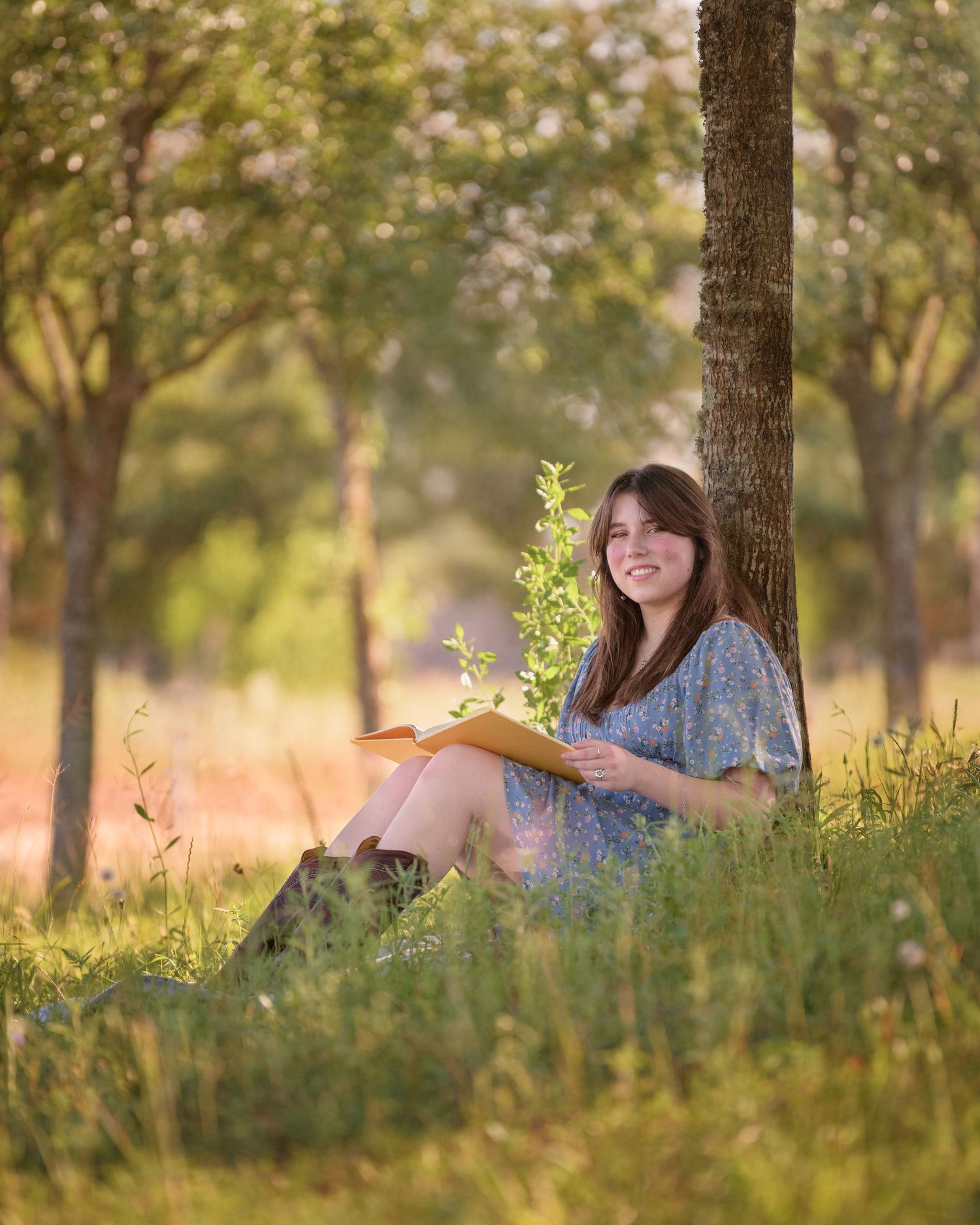 A young woman sits in a grassy meadow at sunset, leaning against a tree trunk in a peaceful natural setting.