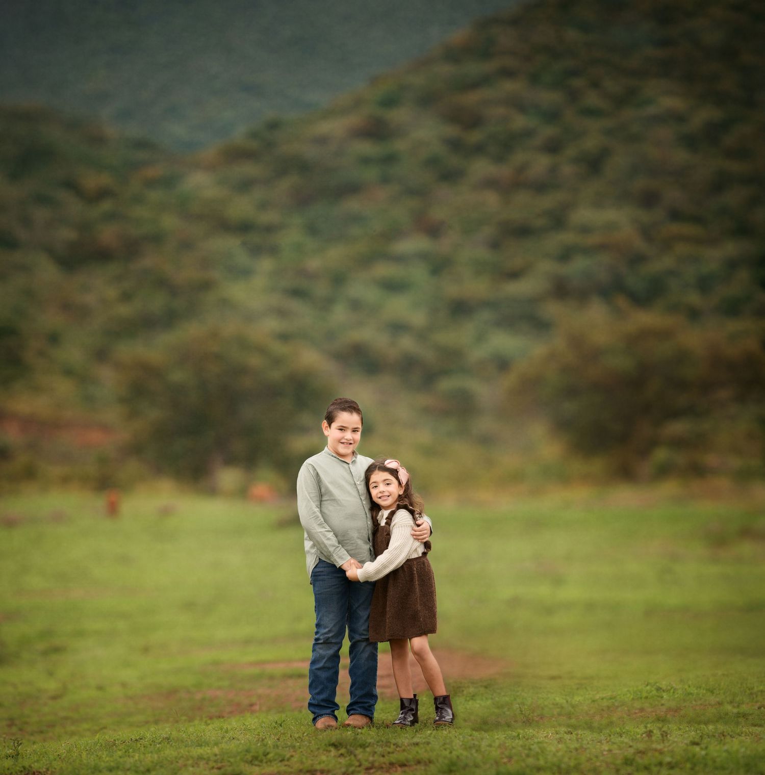 Two kids embrace while standing in green field with mountains in background.