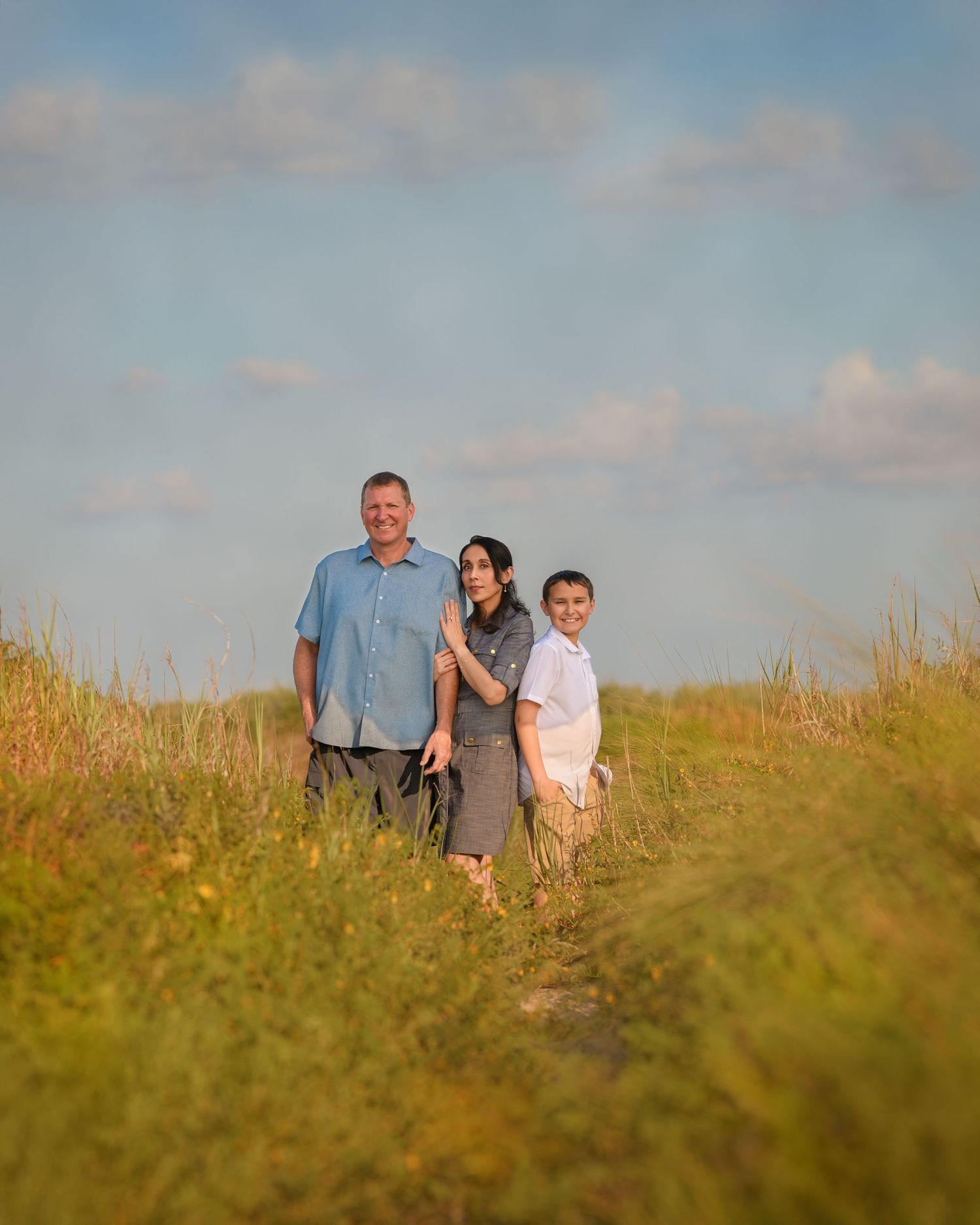 Family of three posing in a sunny green field under a blue sky with fluffy clouds.