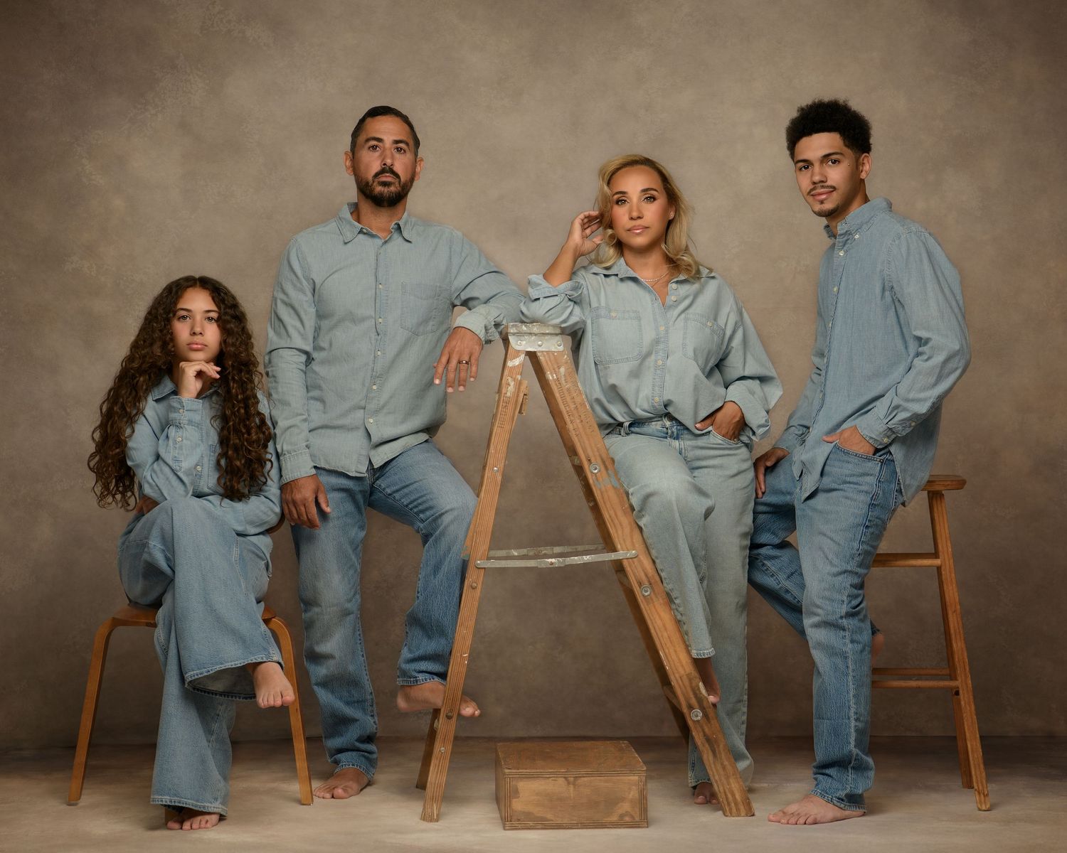 A family wearing matching light blue denim outfits poses together with wooden furniture against a neutral backdrop.