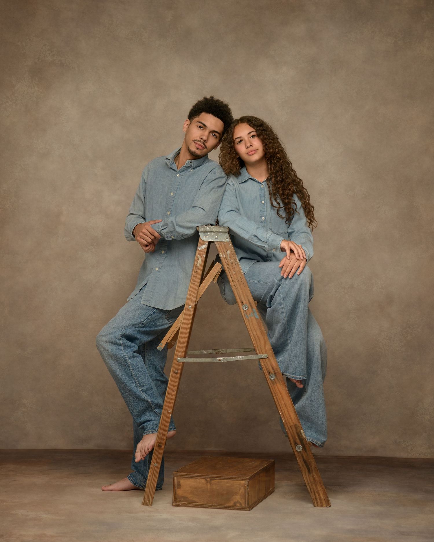 Two people in matching denim outfits sit casually on a wooden ladder against a neutral studio backdrop.